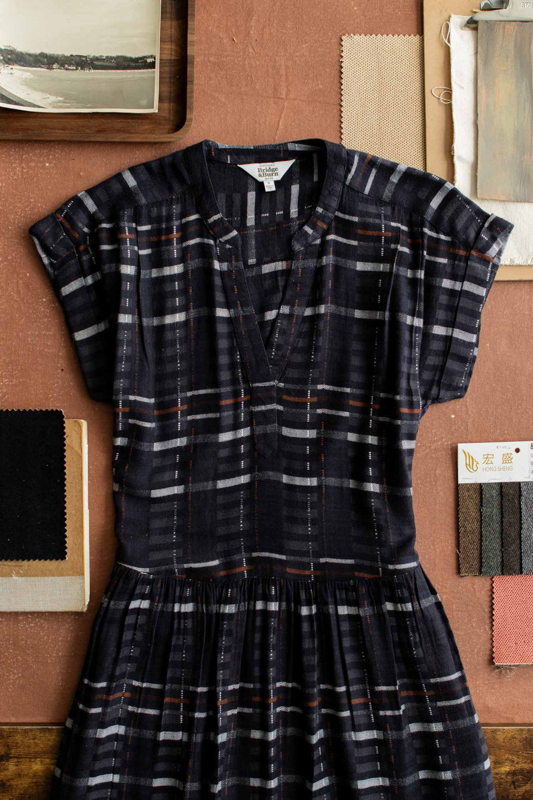 The Bridge & Burn Ira Dress in Nightfall Stripe, a short-sleeve v-neck pullover, is displayed flat on a brown surface with fabric swatches, a notebook, and a painting in the background.