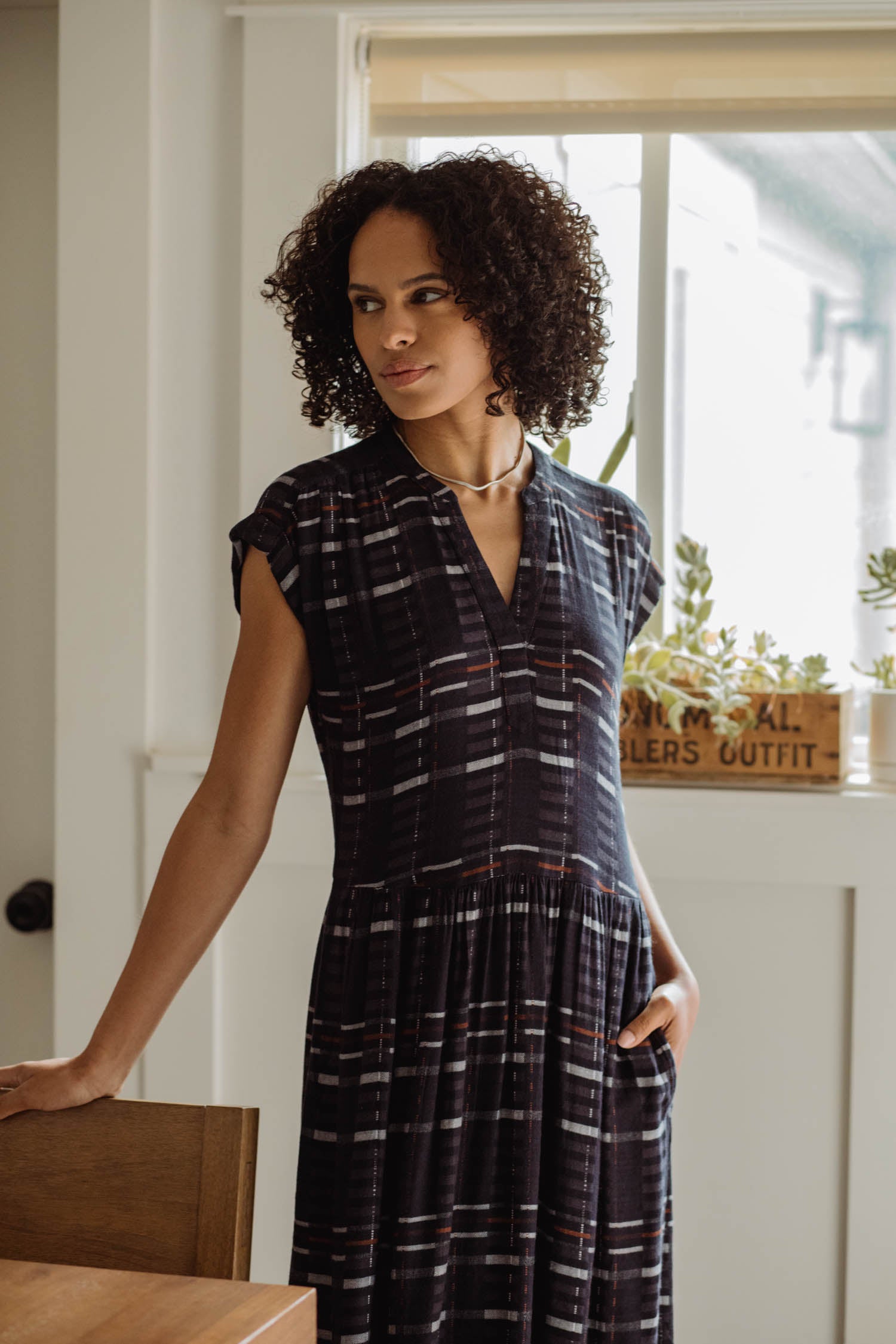 A woman with curly hair stands indoors by a window, wearing the Bridge & Burn Ira Dress in Nightfall Stripe. She looks to the side with one hand on a wooden chair as sunlight streams through behind her.