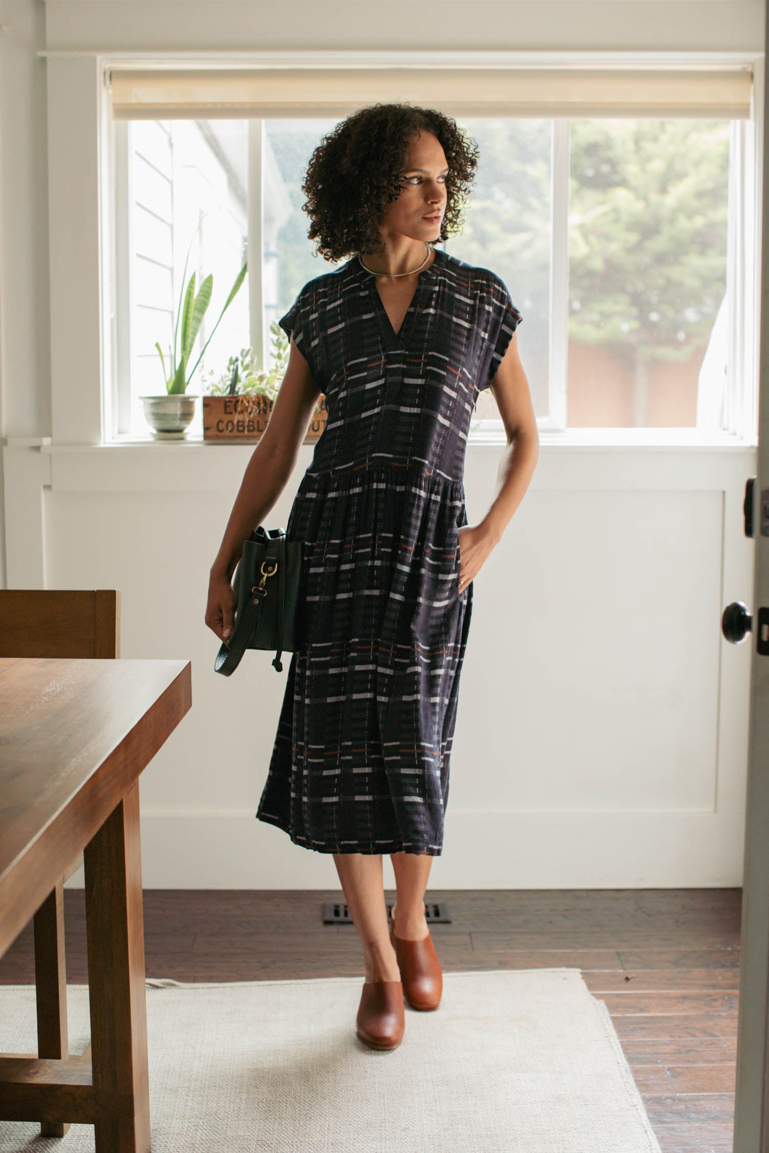 A woman stands indoors near a window, wearing the Bridge & Burn Ira Dress in Nightfall Stripe with brown shoes and holding a black bag. Plants and a sign are visible on the windowsill behind her.