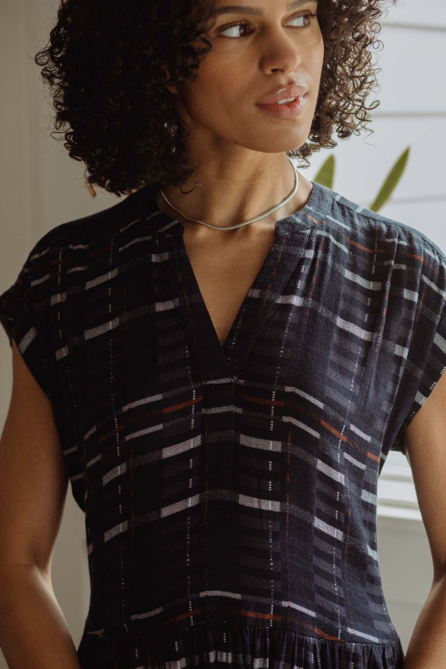 A woman with curly hair stands by a window, wearing the Bridge & Burn Ira Dress in Nightfall Stripe with a silver necklace, gazing calmly to the side.