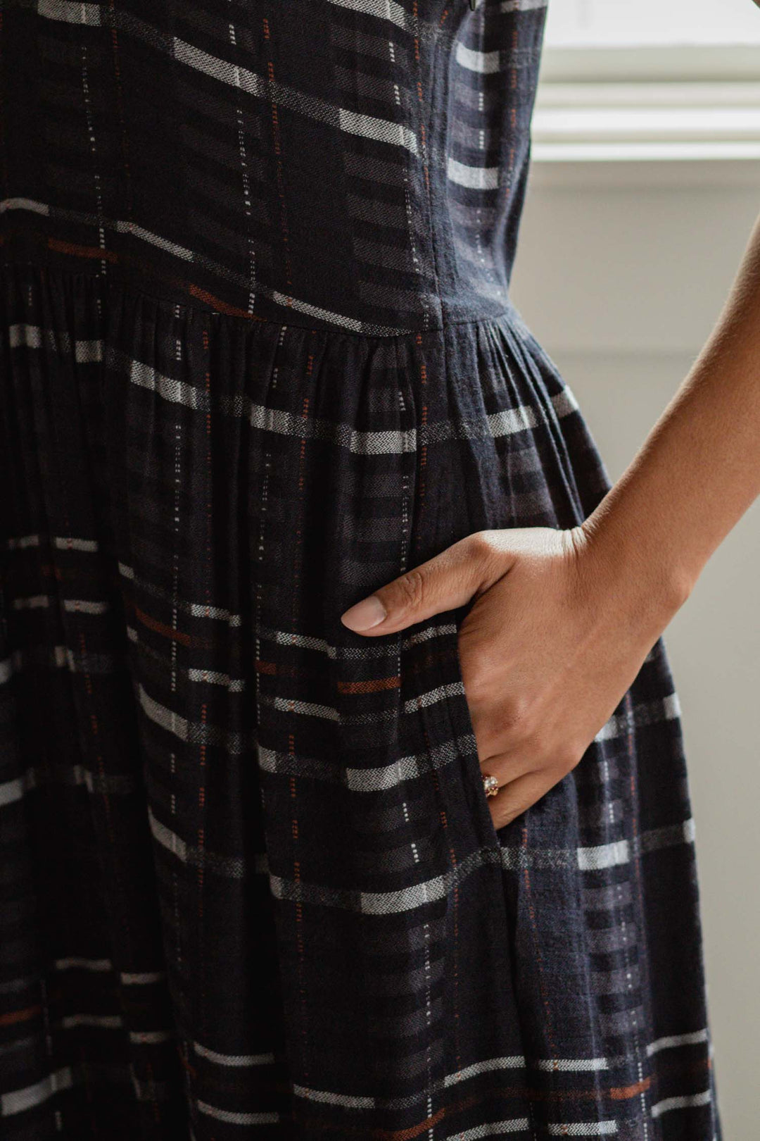 A person’s hand rests in the side pocket of the Bridge & Burn Ira Dress in Nightfall Stripe, a dark pullover dress featuring subtle red, white, and gray stripes. The softly lit backdrop suggests an indoor setting near a window.