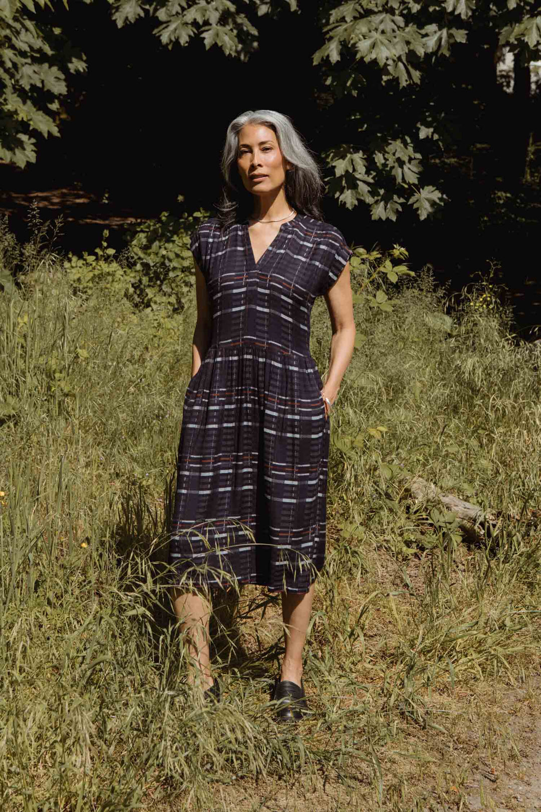A woman with gray hair stands in tall grass, wearing the Bridge & Burn Ira Dress in Nightfall Stripe. The dress features pockets and a gathered skirt, with trees and sunlight visible in the background.