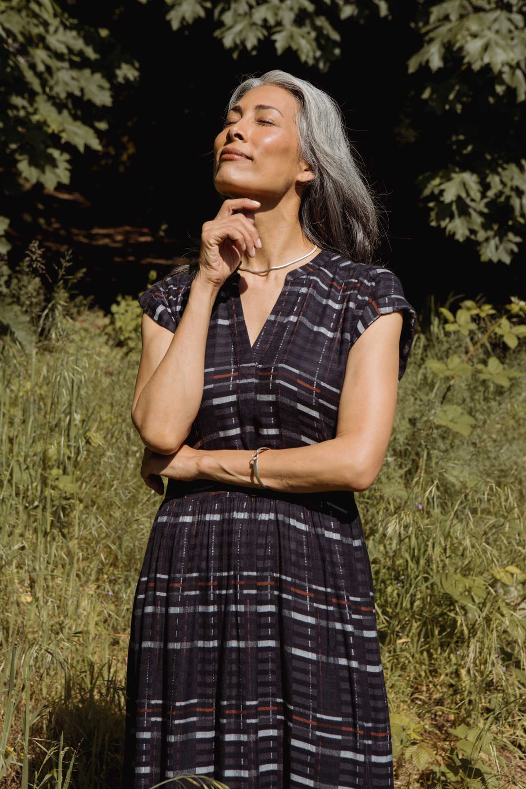 A woman with long gray hair stands outside in sunlight, wearing the Bridge & Burn Ira Dress in Nightfall Stripe. She crosses one arm and rests her hand on her chin, surrounded by green foliage and tall grass.