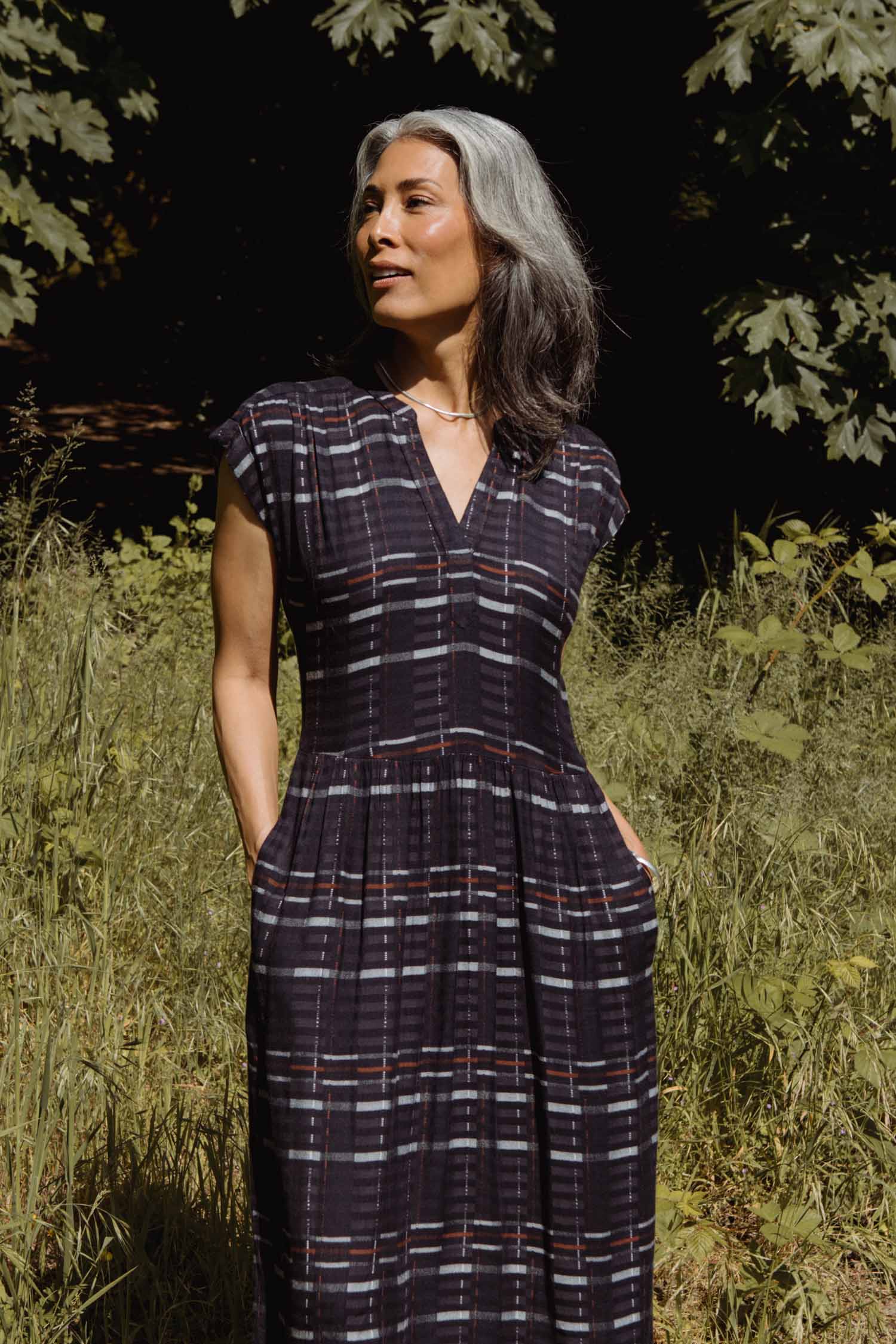 A woman with long, gray-streaked hair stands outdoors among tall grass and leafy trees, wearing the Bridge & Burn Ira Dress in Nightfall Stripe, hands in pockets, looking to the side and smiling gently.