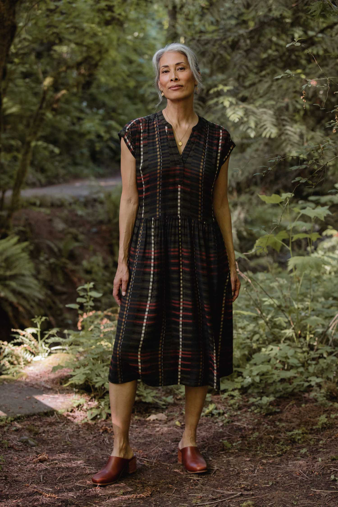 A woman with grey hair in a low bun stands on a wooded path, surrounded by lush green foliage, wearing the Bridge & Burn Ira Dress in Olive Stripe—a knee-length, wrinkle-resistant pullover dress—paired with brown shoes.