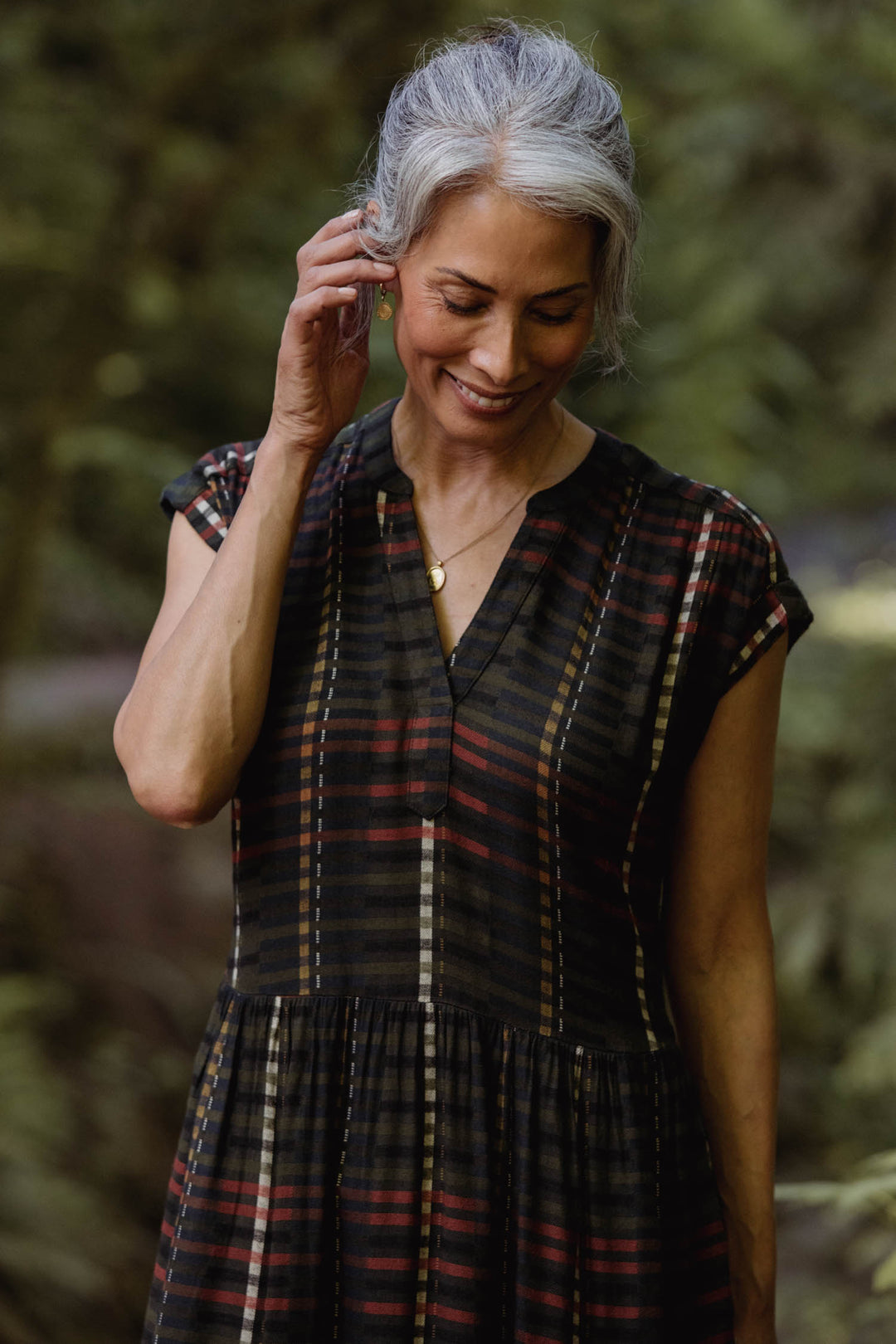 A woman with gray hair wears the Bridge & Burn Ira Dress in Olive Stripe, standing outdoors and gently touching her ear as she smiles, with blurred greenery in the background.