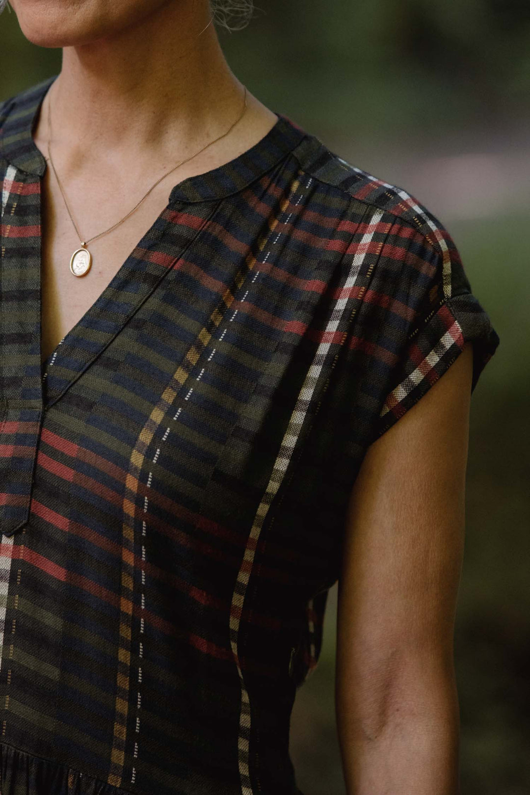 Someone wearing the Bridge & Burn Ira Dress in Olive Stripe, a short-sleeved, wrinkle-resistant pullover with stripes, paired with a gold pendant necklace. The cropped photo shows the upper torso and neck against a blurred outdoor background.