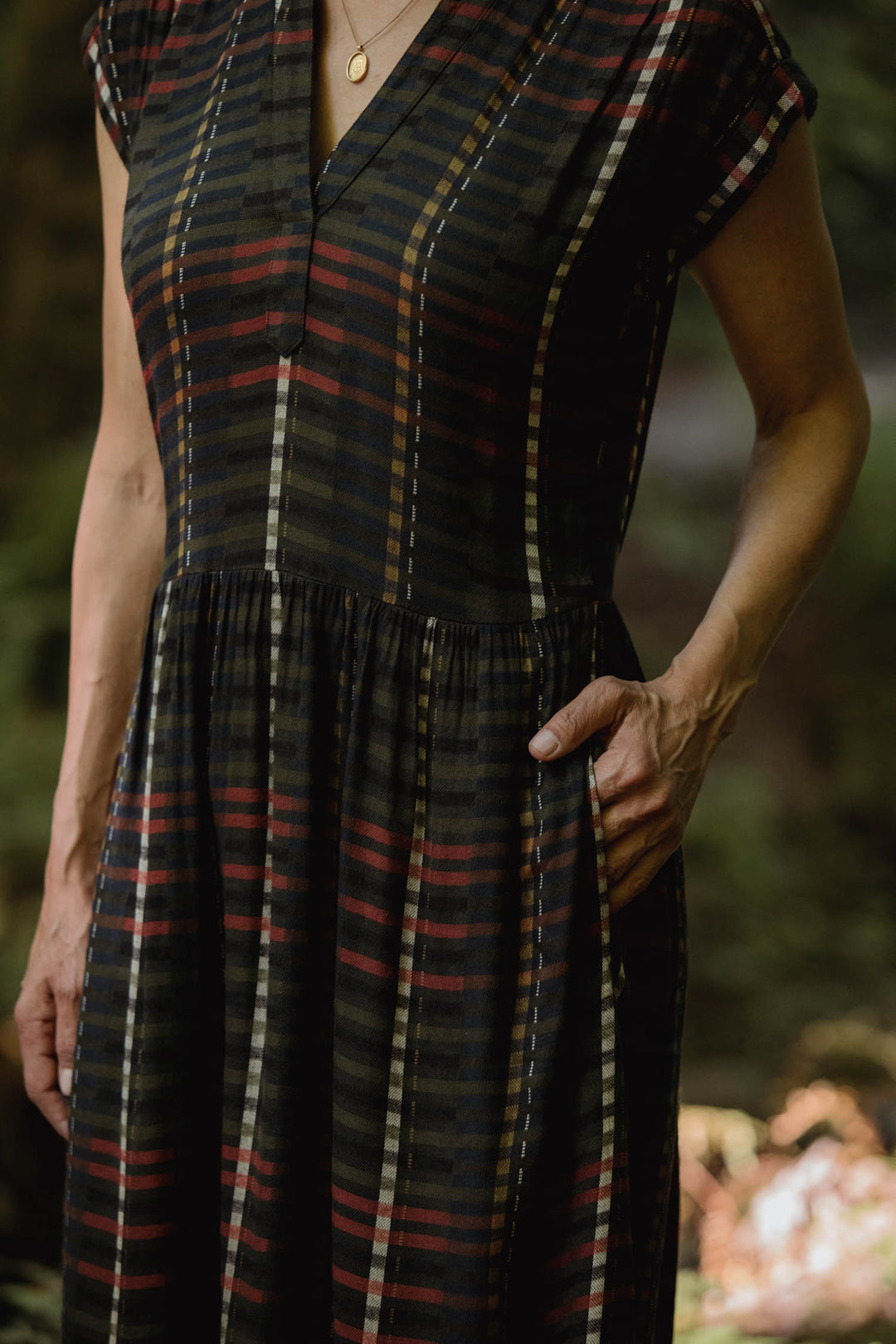 A person stands outdoors with one hand in their pocket, wearing the Bridge & Burn Ira Dress in Olive Stripe. The dress is short-sleeved, wrinkle-resistant, and features multi-colored stripes. Their head is not visible in the image.