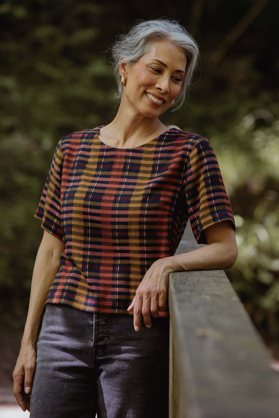 A woman with gray hair smiles while leaning on a wooden railing outdoors, wearing the Bridge & Burn Leona Shirt in Alpenglow Stripe, boxy fit and made from eco-friendly viscose. Blurred green foliage fills the background.