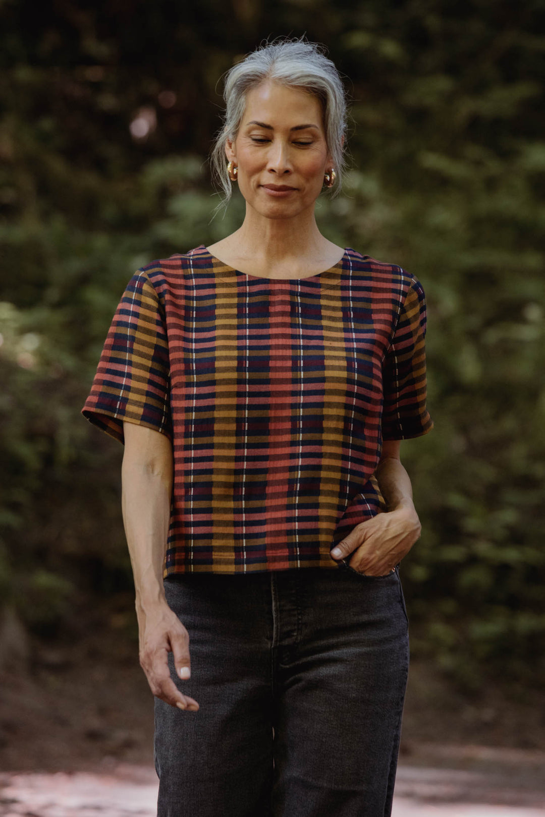 A woman with gray hair in a ponytail wears the Bridge & Burn Leona Shirt in Alpenglow Stripe and dark jeans, standing outdoors among trees with one hand in her pocket and a calm expression.