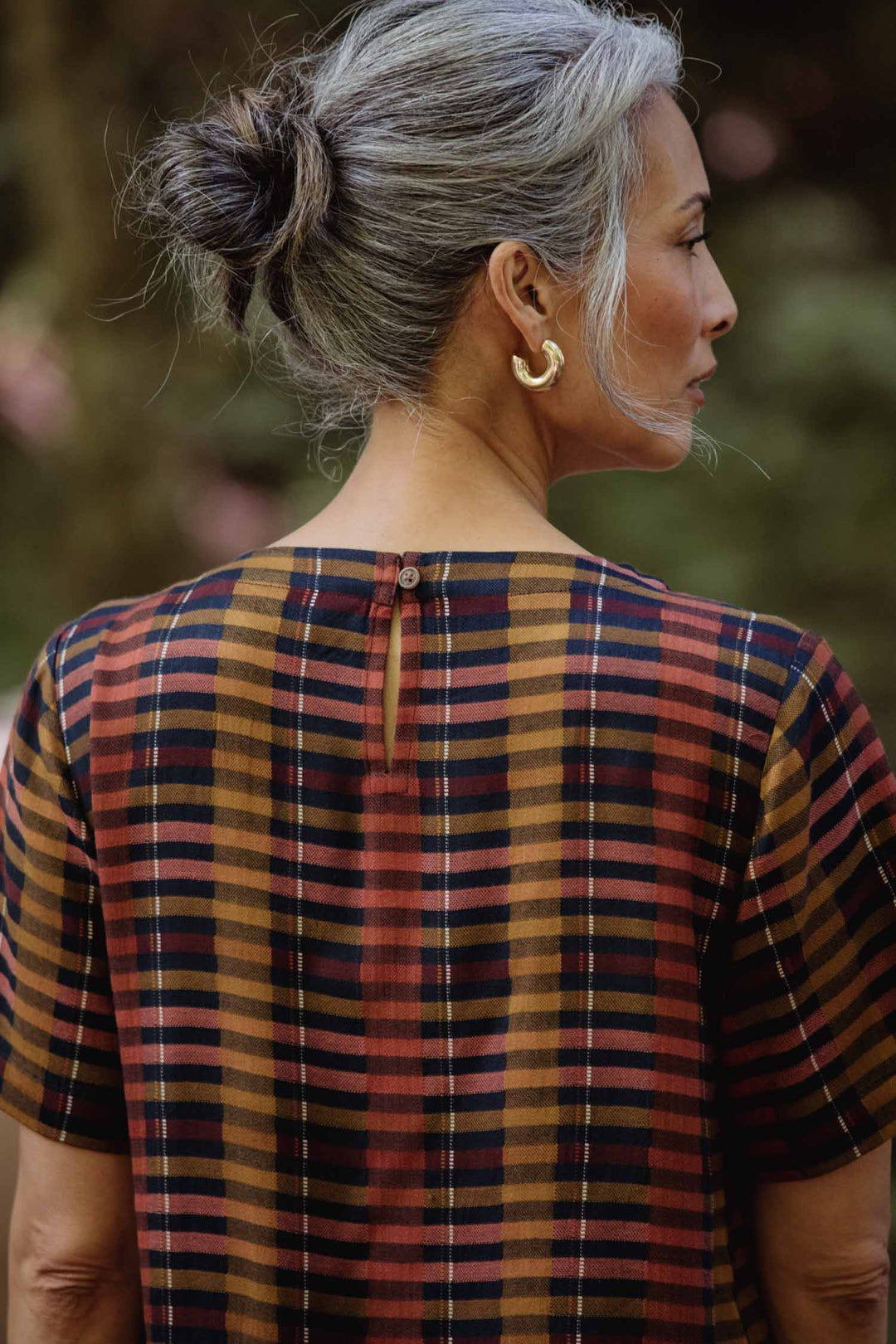 A woman with gray hair in a bun and gold hoop earrings wears the Bridge & Burn Leona Shirt in Alpenglow Stripe, a short-sleeved boxy blouse with a cropped hem, shown from the back and side against a blurred outdoor background.