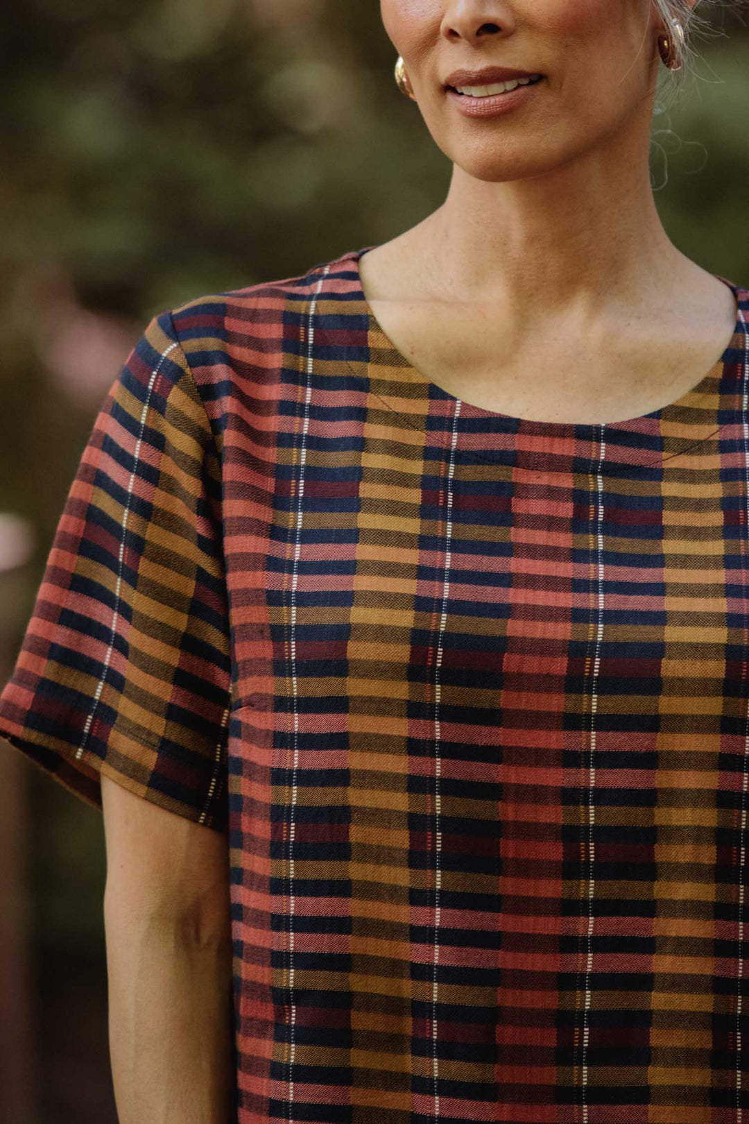 A woman wears the Bridge & Burn Leona Shirt in Alpenglow Stripe, a boxy blouse with short sleeves and a cropped hem, featuring multicolored stripes. Made from eco-friendly viscose, she stands outdoors with only her upper body visible.