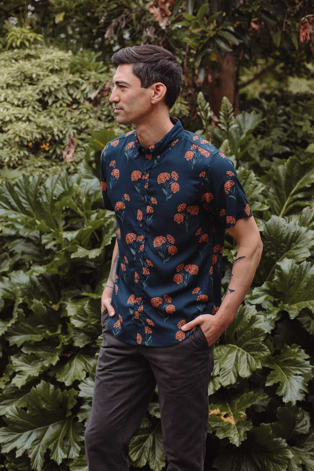 A man stands outdoors before lush foliage, wearing the Bridge & Burn Grant Slim Shirt in Wildflower print and dark pants, with his hands in his pockets, looking to the left.