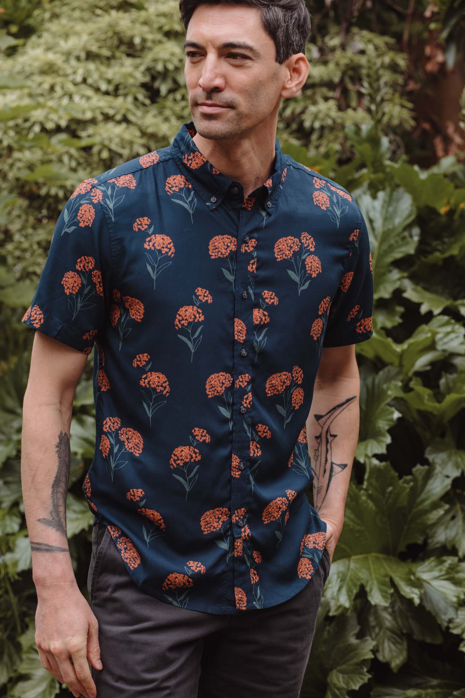 A man with tattooed forearms stands outdoors by green foliage, wearing the Bridge & Burn Grant Slim Shirt / Wildflower—a navy blue slim fit shirt with a red floral pattern—and dark pants, looking slightly to the side.