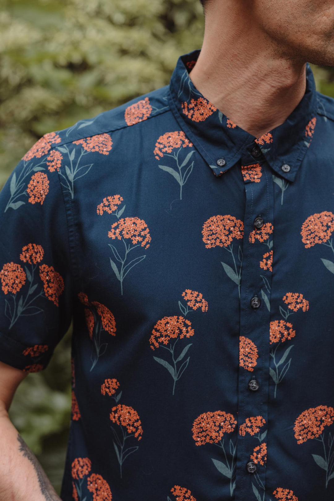 A man wears the Bridge & Burn Grant Slim Shirt / Wildflower—a navy blue, slim-fit modal shirt with orange floral pattern—standing out against blurred greenery, his face partially out of frame.