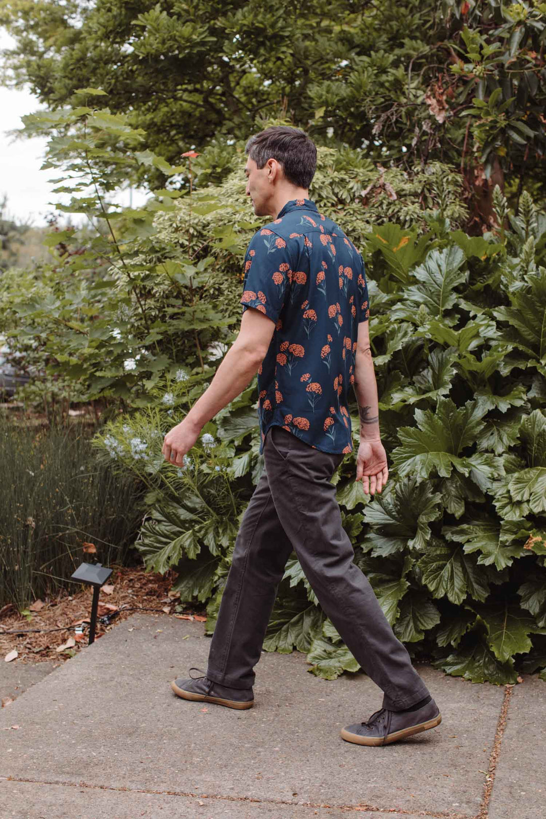 A man wearing the Bridge & Burn Grant Slim Shirt in Wildflower and dark pants walks on a concrete path surrounded by lush green plants and trees.