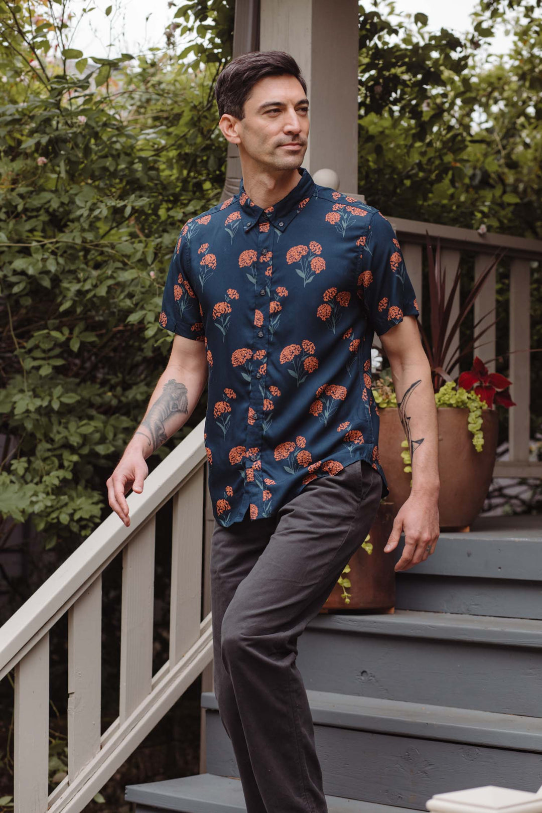 A man with short dark hair, in a Bridge & Burn Grant Slim Shirt / Wildflower and dark pants, walks down wooden steps outside among green plants and potted flowers.