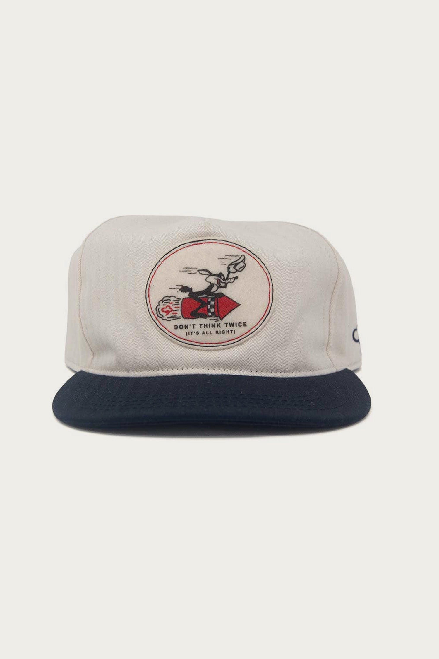 The Ampal Creative - Don't Think Twice Snapback / Navy by The Ampal Creative is a cream-colored hat with a navy brim, featuring an Ampal patch of a person on a red motorcycle and "Don't Think Twice (It's All Right)" text, nodding to Bob Dylan’s song.