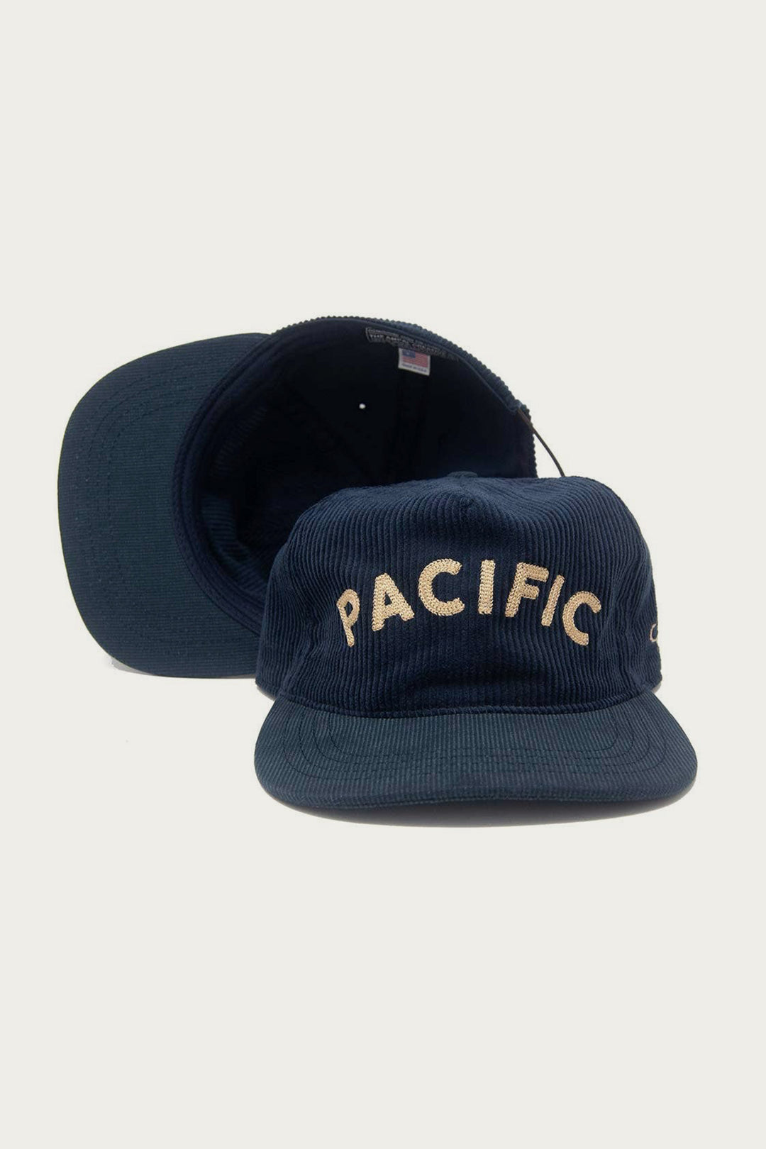 Two navy blue The Ampal Creative - Pacific III Strapback hats are shown: one upright with “PACIFIC” in cream vintage-style embroidery, the other lying flat behind—a must-have for classic style fans.