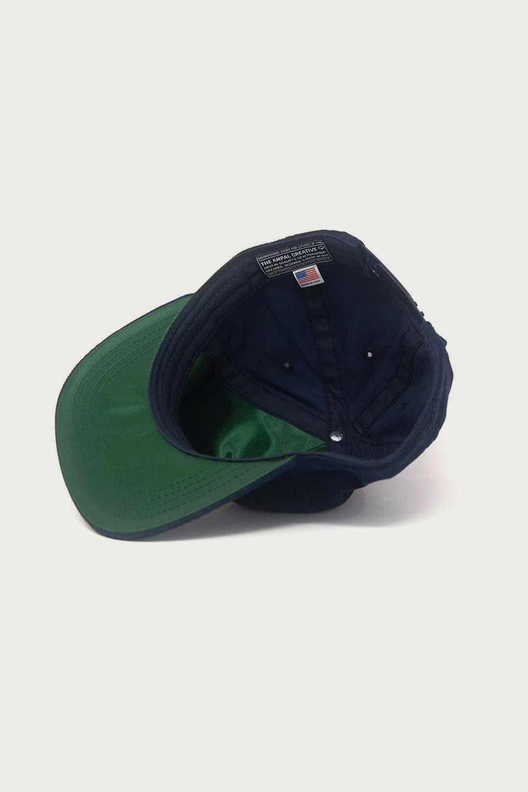 The Ampal Creative - Tiger III Snapback / Navy is shown upside down on a white background, displaying its green underbrim and an American flag label sewn inside the crown. Made in Los Angeles by The Ampal Creative.