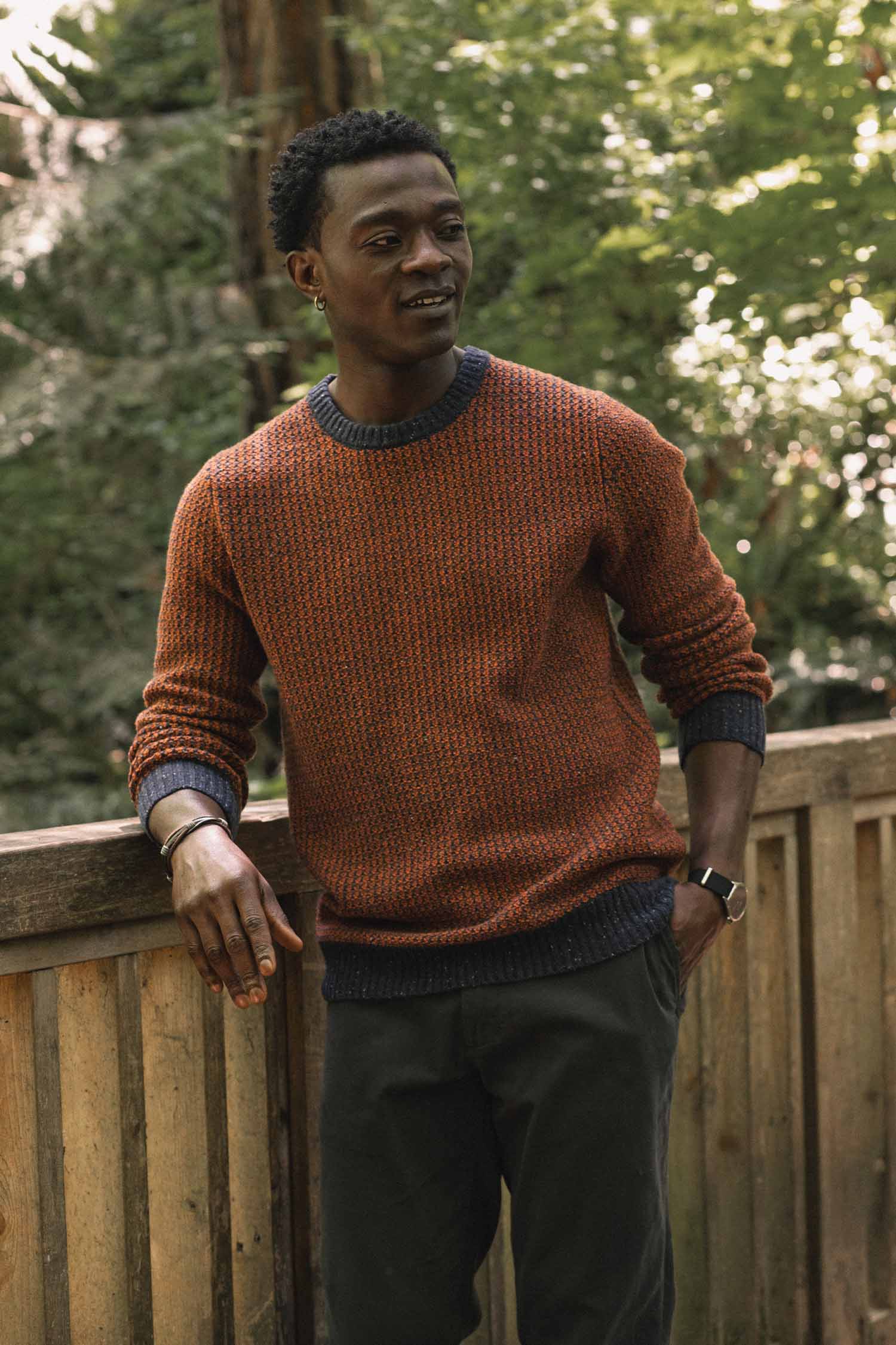 A man wearing the Bridge & Burn Brighton Sweater in Autumn Tide stands by a wooden railing outdoors, surrounded by lush green trees and sunlight.