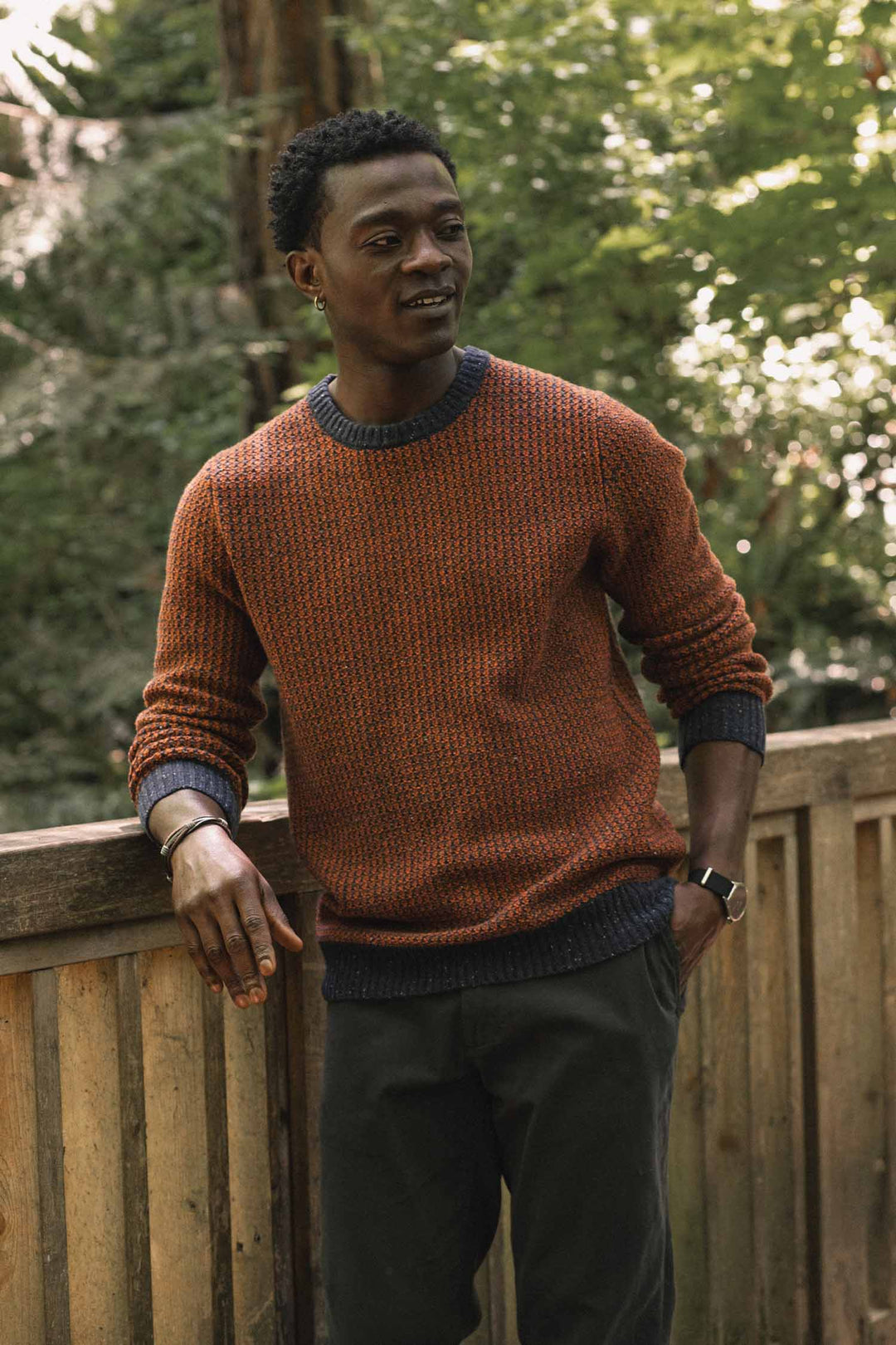 A man wearing the Bridge & Burn Brighton Sweater in Autumn Tide stands by a wooden railing outdoors, surrounded by lush green trees and sunlight.
