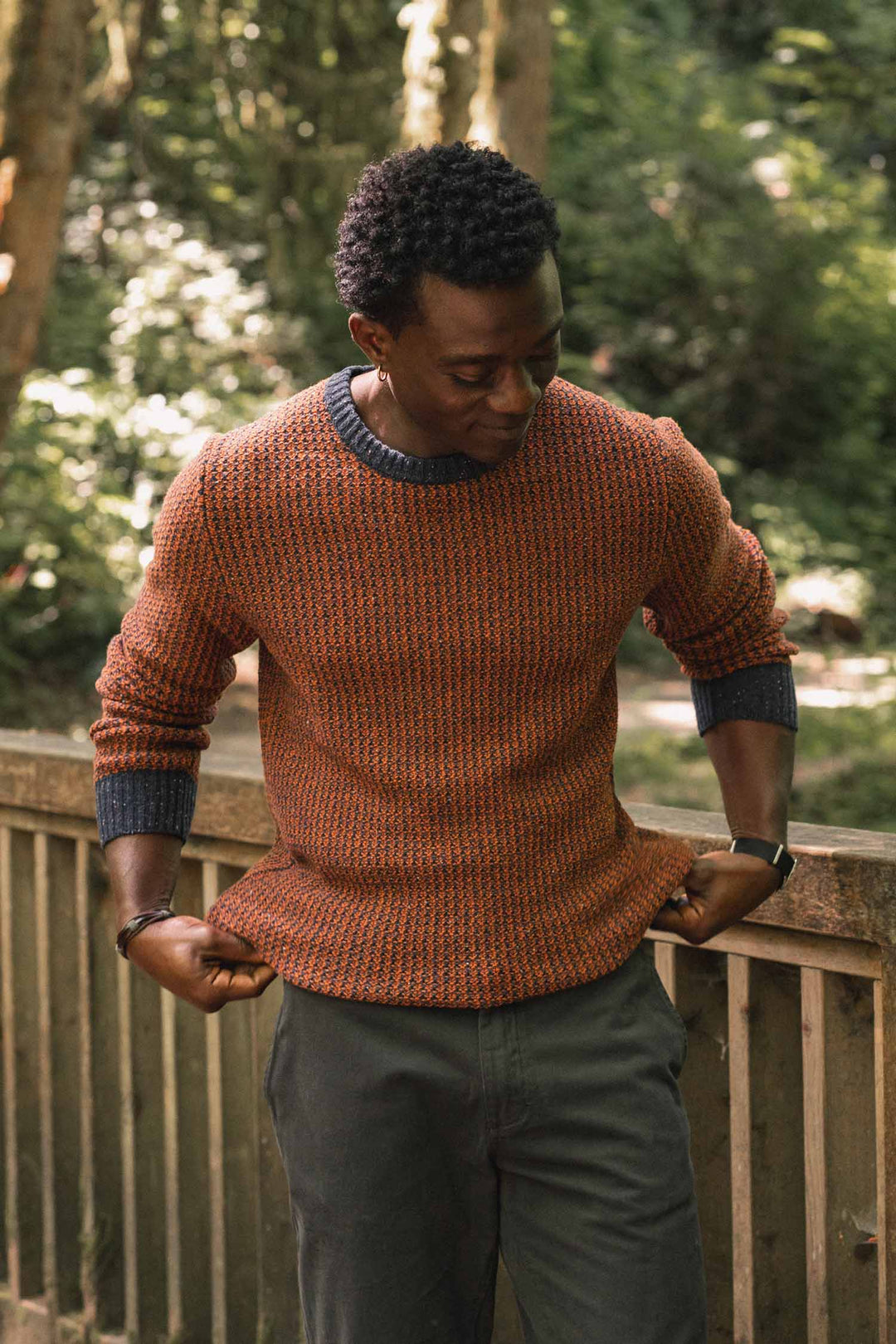 A man wearing the Bridge & Burn Brighton Sweater in Autumn Tide stands outdoors near a wooden railing, looking down and slightly tugging at his sweater. Trees and greenery fill the background.