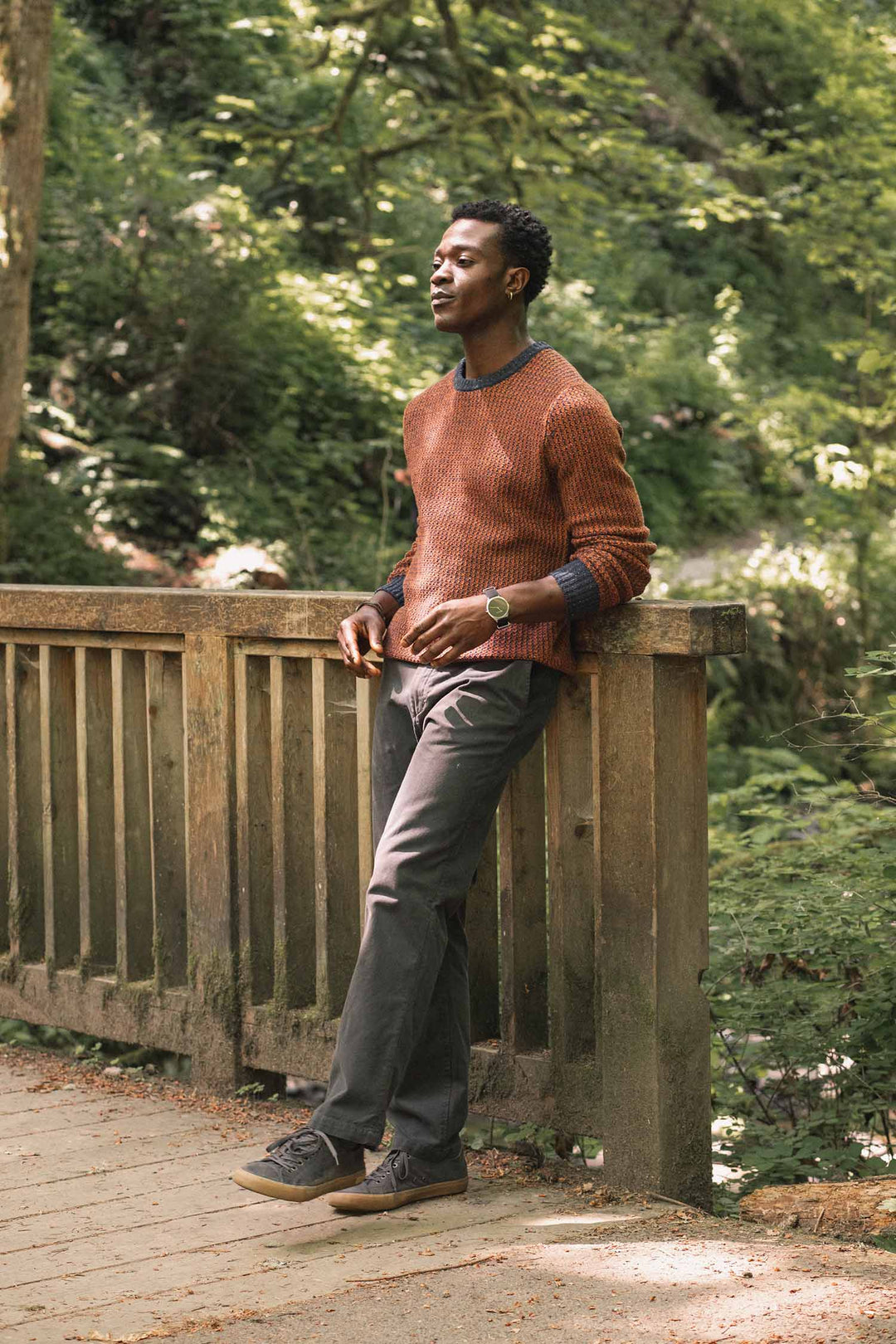 A man wearing the Bridge & Burn Brighton Sweater in Autumn Tide and dark pants leans casually on a wooden bridge in a lush, green forest, enjoying the natural surroundings.