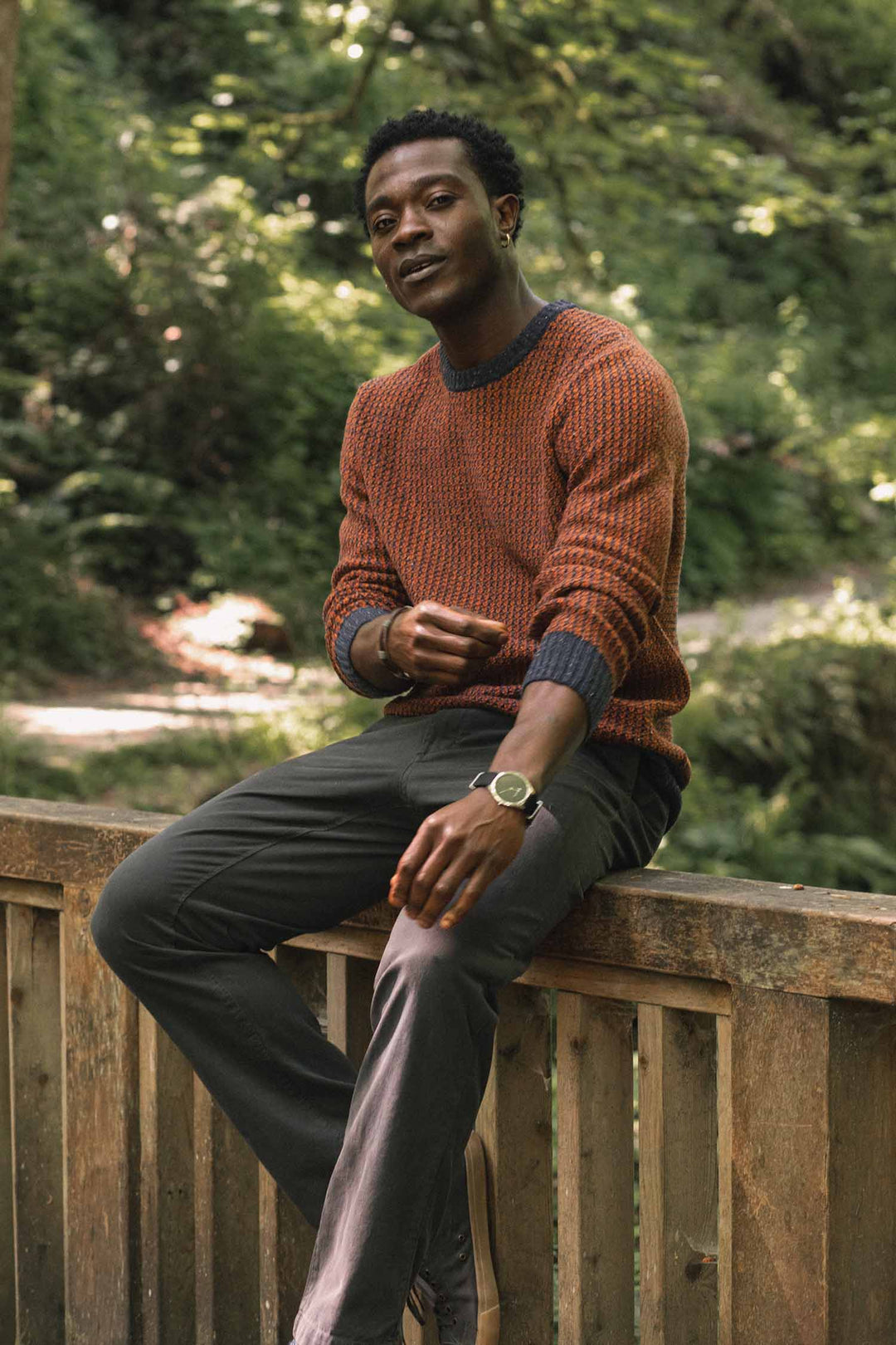 A man wearing the Bridge & Burn Brighton Sweater in Autumn Tide and dark pants sits on a wooden railing outdoors, surrounded by green trees, looking relaxed and confident.