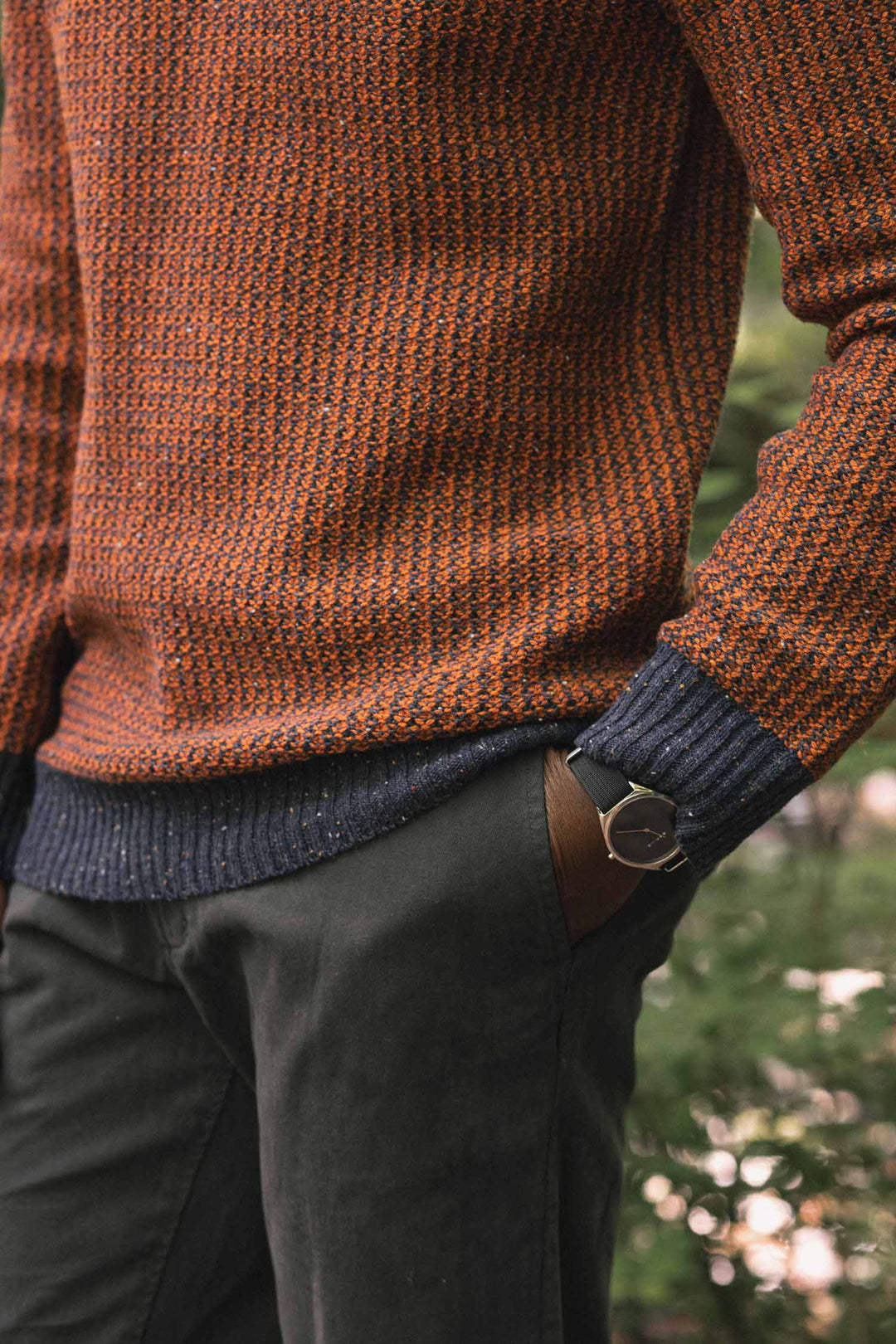 A person wears the Bridge & Burn Brighton Sweater in Autumn Tide, paired with olive green pants and a wristwatch, standing outdoors with one hand in their pocket.
