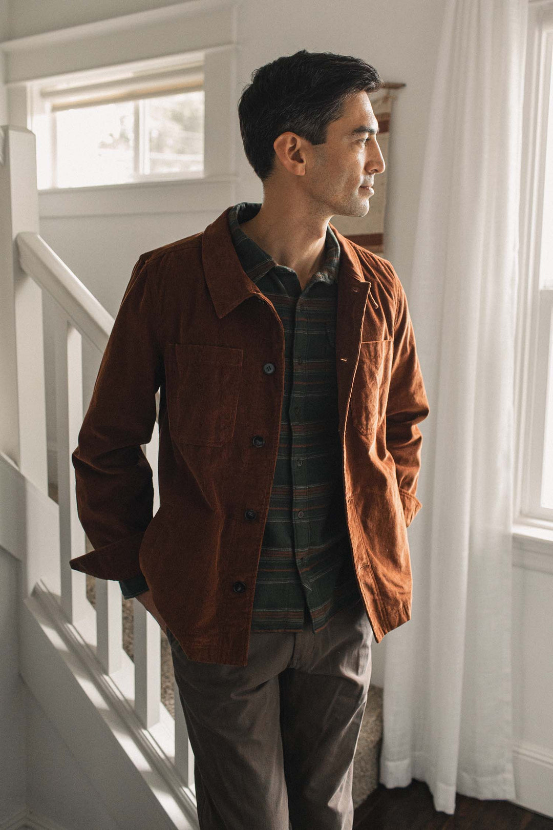 A man stands indoors near a staircase, looking out a sunlit window while wearing the Bridge & Burn Banks Shirt Jacket in Saddle Brown over a striped shirt and brown pants.