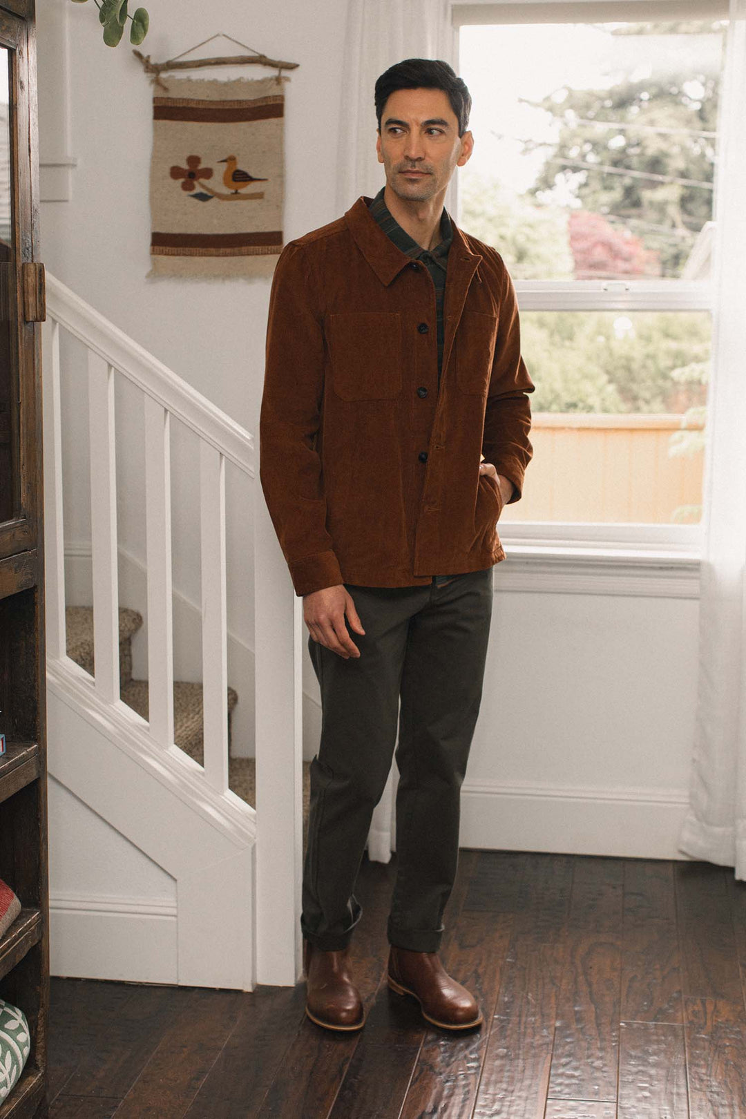 A man stands indoors by a staircase and window, wearing the Bridge & Burn Banks Shirt Jacket in Saddle Brown, dark pants, and brown boots. The room has wooden floors, white walls, and a small bird-themed wall hanging.