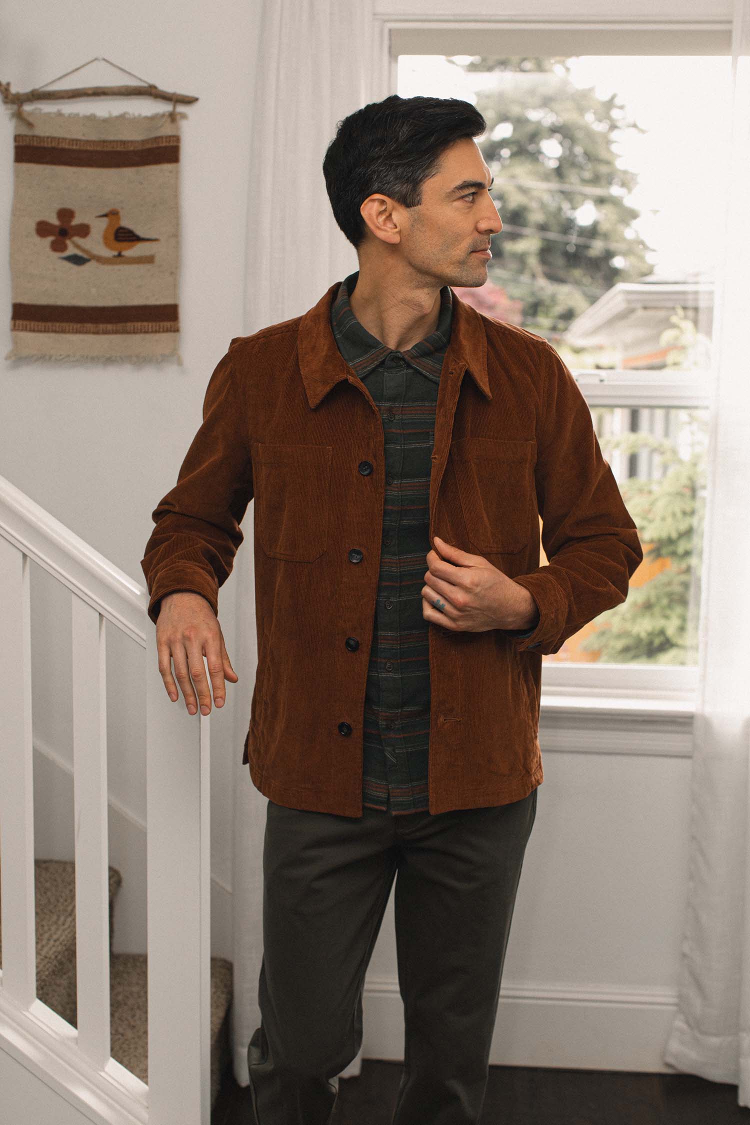 A man stands indoors near a staircase, wearing the Bridge & Burn Banks Shirt Jacket in Saddle Brown over a plaid shirt and dark pants. He looks to the side, with a window and decorative wall hanging in the background.