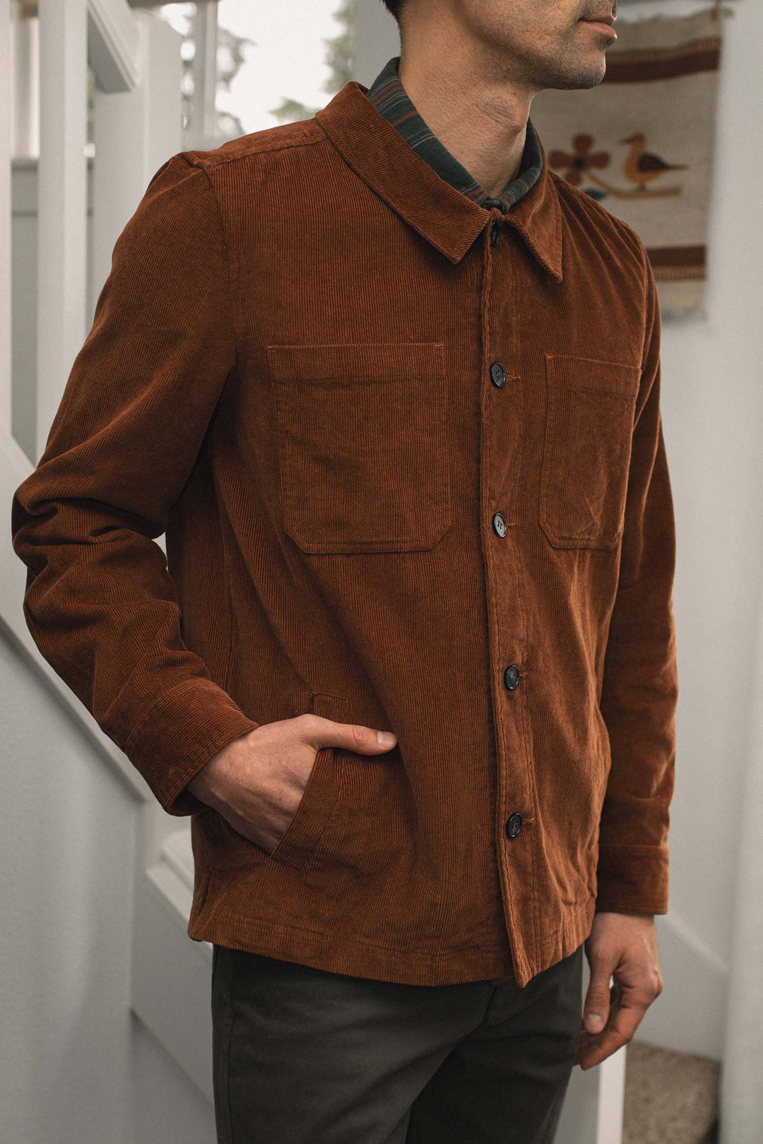 A man wears the Bridge & Burn Banks Shirt Jacket in Saddle Brown, featuring two chest pockets, standing indoors with one hand in his pocket. His head is partially out of frame, with white stairs and a patterned wall hanging behind him.