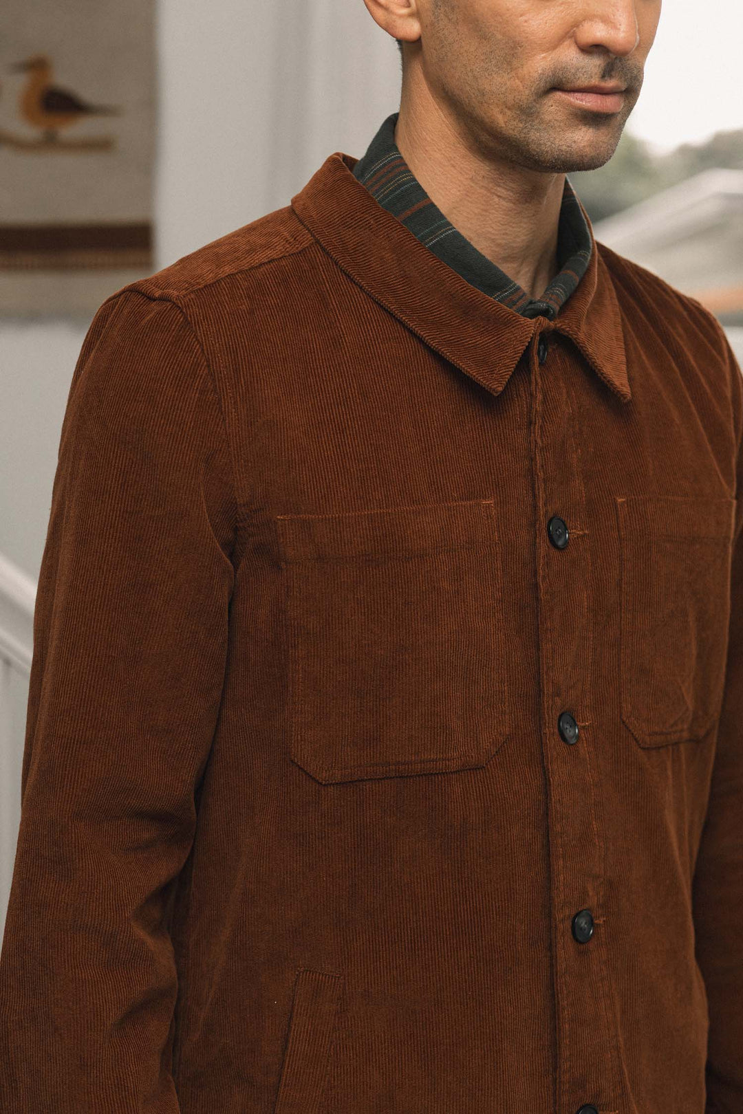 A man wearing the Bridge & Burn Banks Shirt Jacket in Saddle Brown, layered over a collared shirt, stands indoors with his face partially out of frame. The jacket features large front pockets and a corduroy texture.