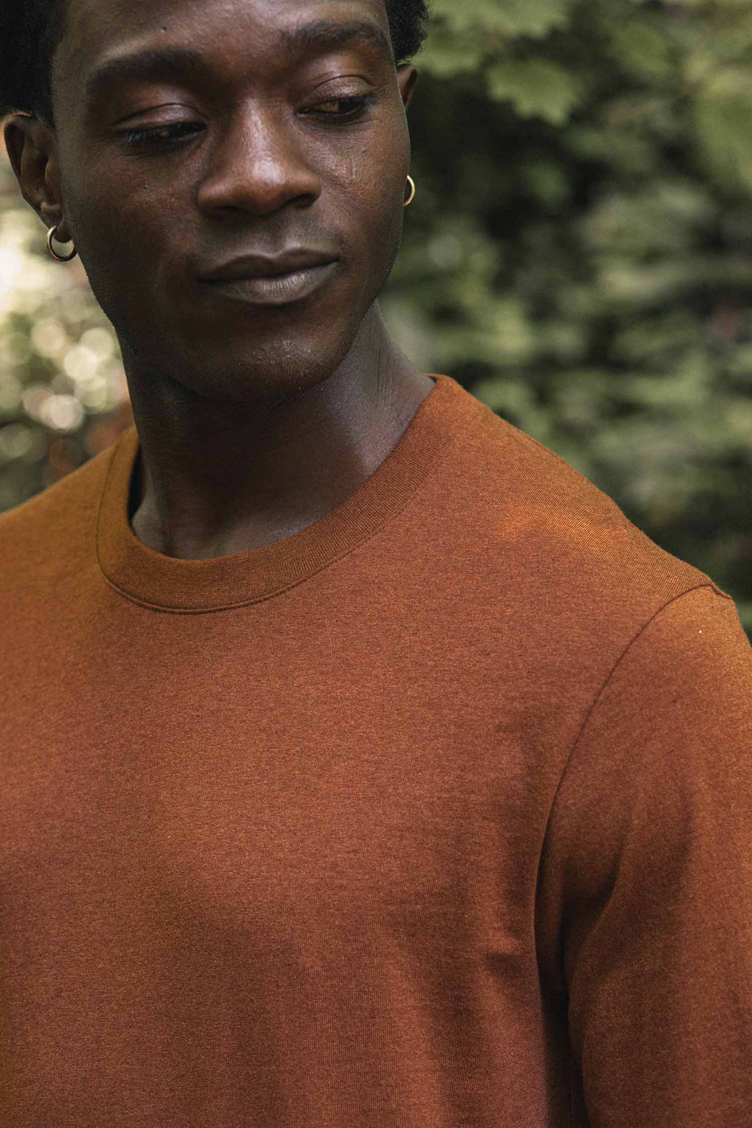 Wearing the Bridge & Burn Barney Long Sleeve Tee in Bronze and hoop earrings, a person looks down while standing outdoors with blurred greenery in the background.