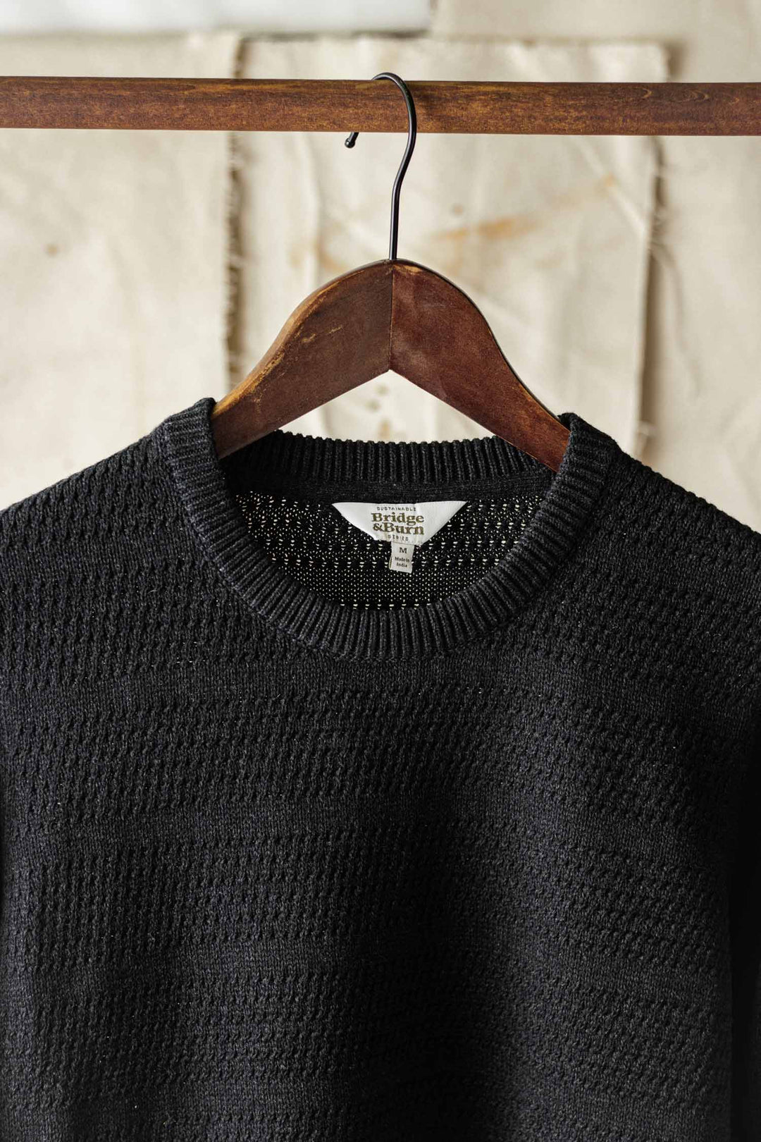 The Bridge & Burn Calder Sweater in Midnight, featuring a black textured sustainable design, hangs on a wooden hanger against a neutral, slightly wrinkled background.
