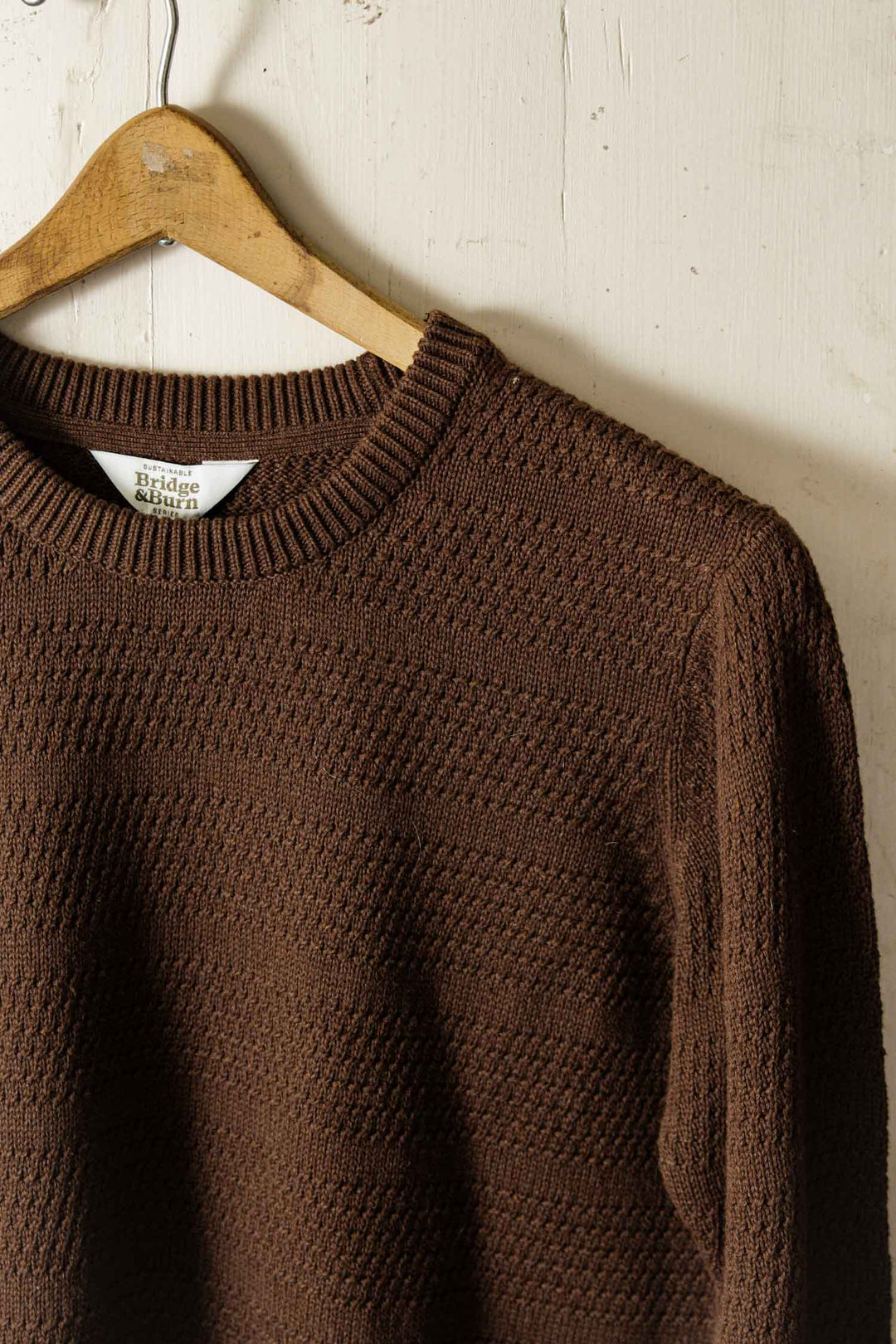 A close-up of the Bridge & Burn Calder Sweater in Walnut shows its textured knit and ribbed neckline, hanging on a wooden hanger against a light wood wall—a sustainable, stylish choice.