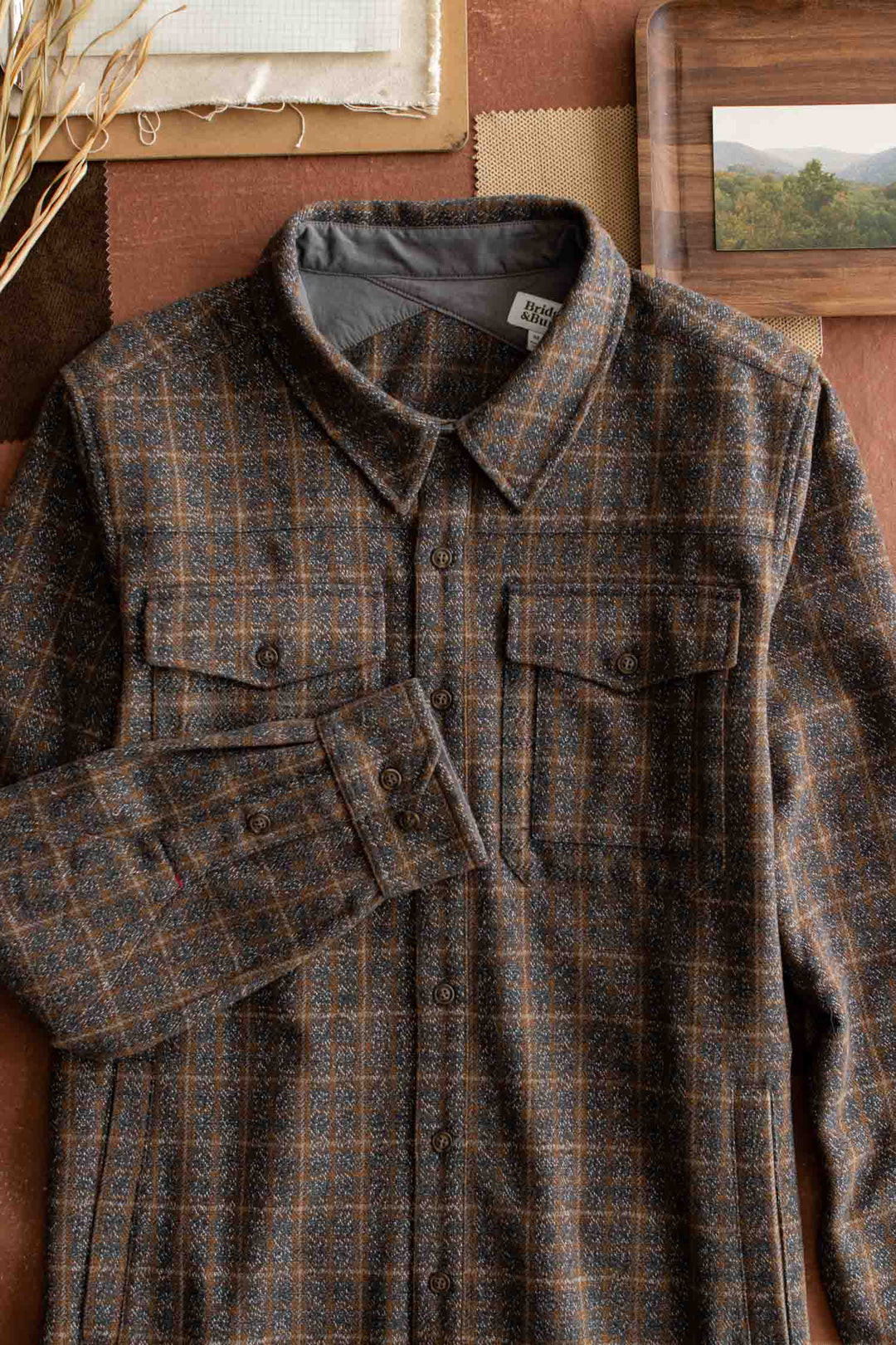 The Bridge & Burn Fielding Shirt Jacket in Hearth Plaid is laid flat on a brown surface, surrounded by fabric swatches, dried grass, and a framed landscape photo.
