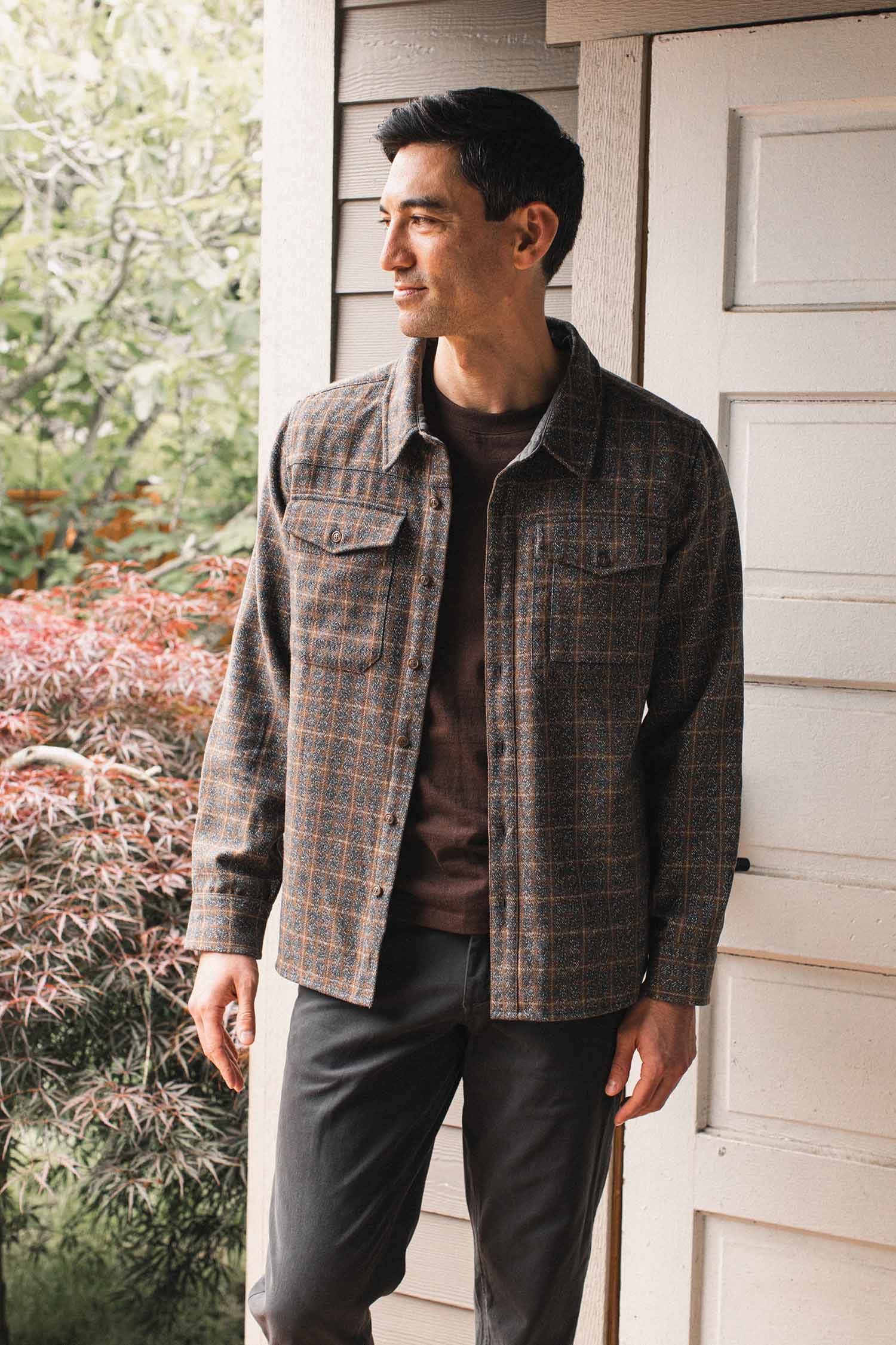 A man stands outside by a light wood wall and door, wearing the Bridge & Burn Fielding Shirt Jacket in Hearth Plaid over dark clothes, looking to his left. Greenery and a reddish shrub are visible in the background.
