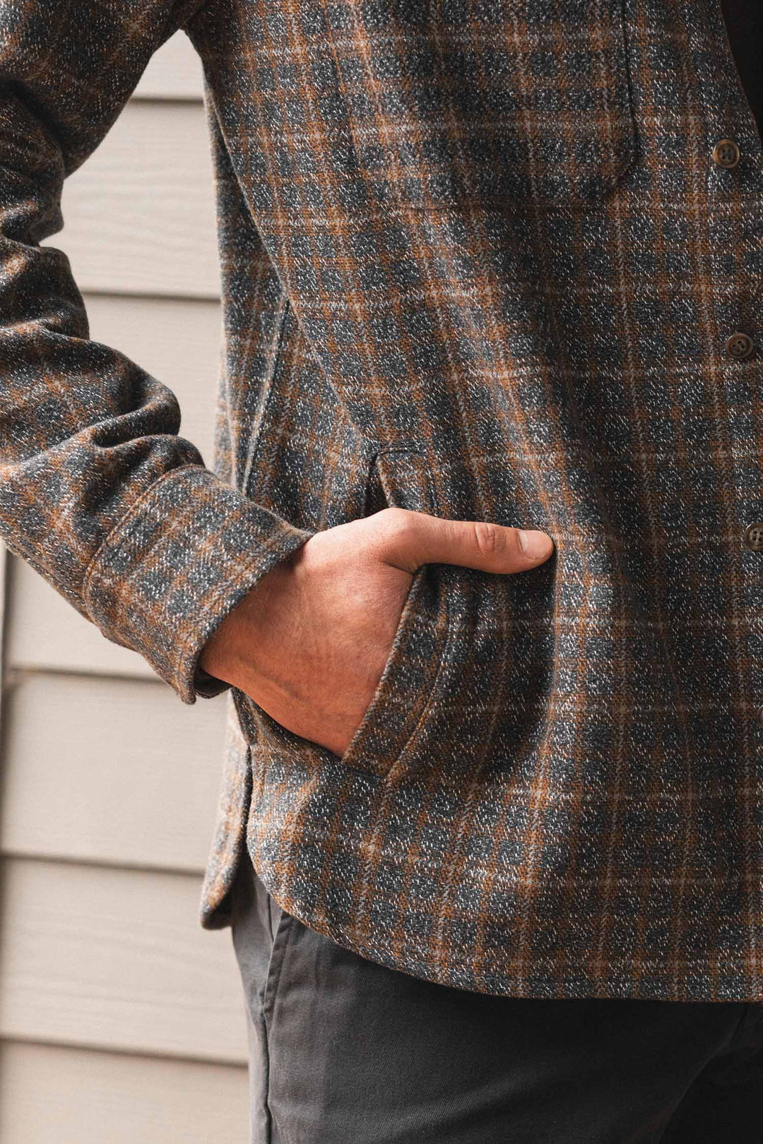 A person wearing the Bridge & Burn Fielding Shirt Jacket in Hearth Plaid stands sideways with their right hand in the pocket, next to a wall with horizontal beige siding.