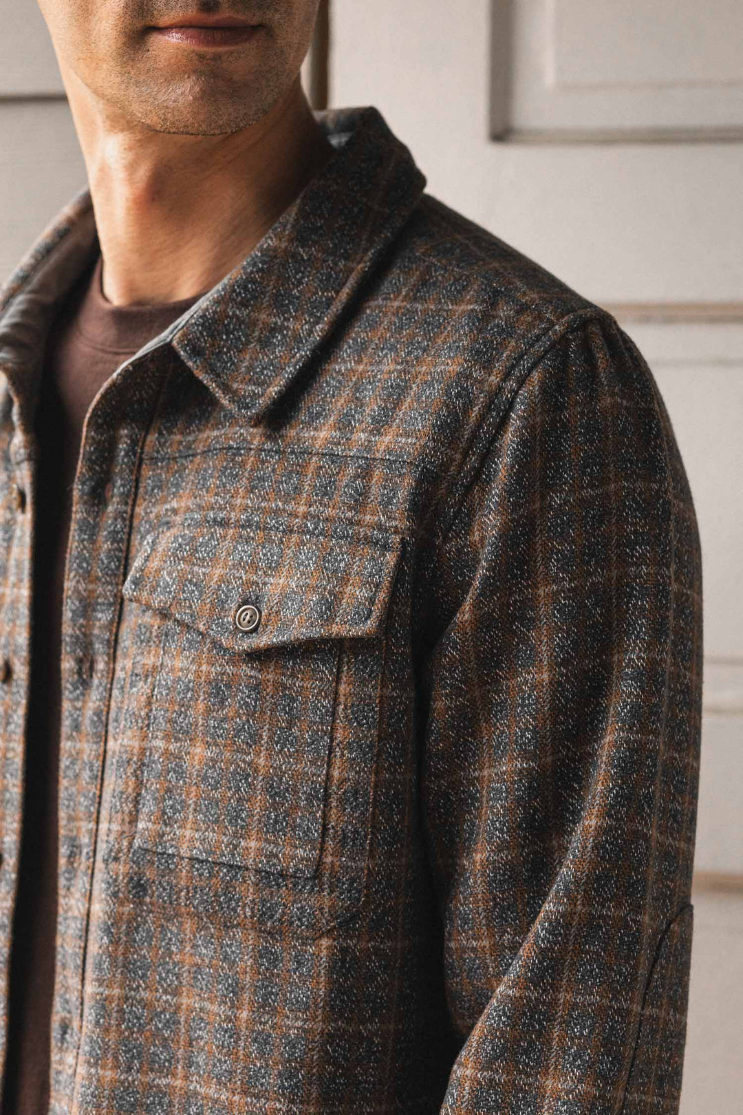A man wearing the Bridge & Burn Fielding Shirt Jacket in Hearth Plaid stands indoors, partially facing the camera. The photo highlights the shirt's texture and chest pocket against a softly blurred, light-paneled background.