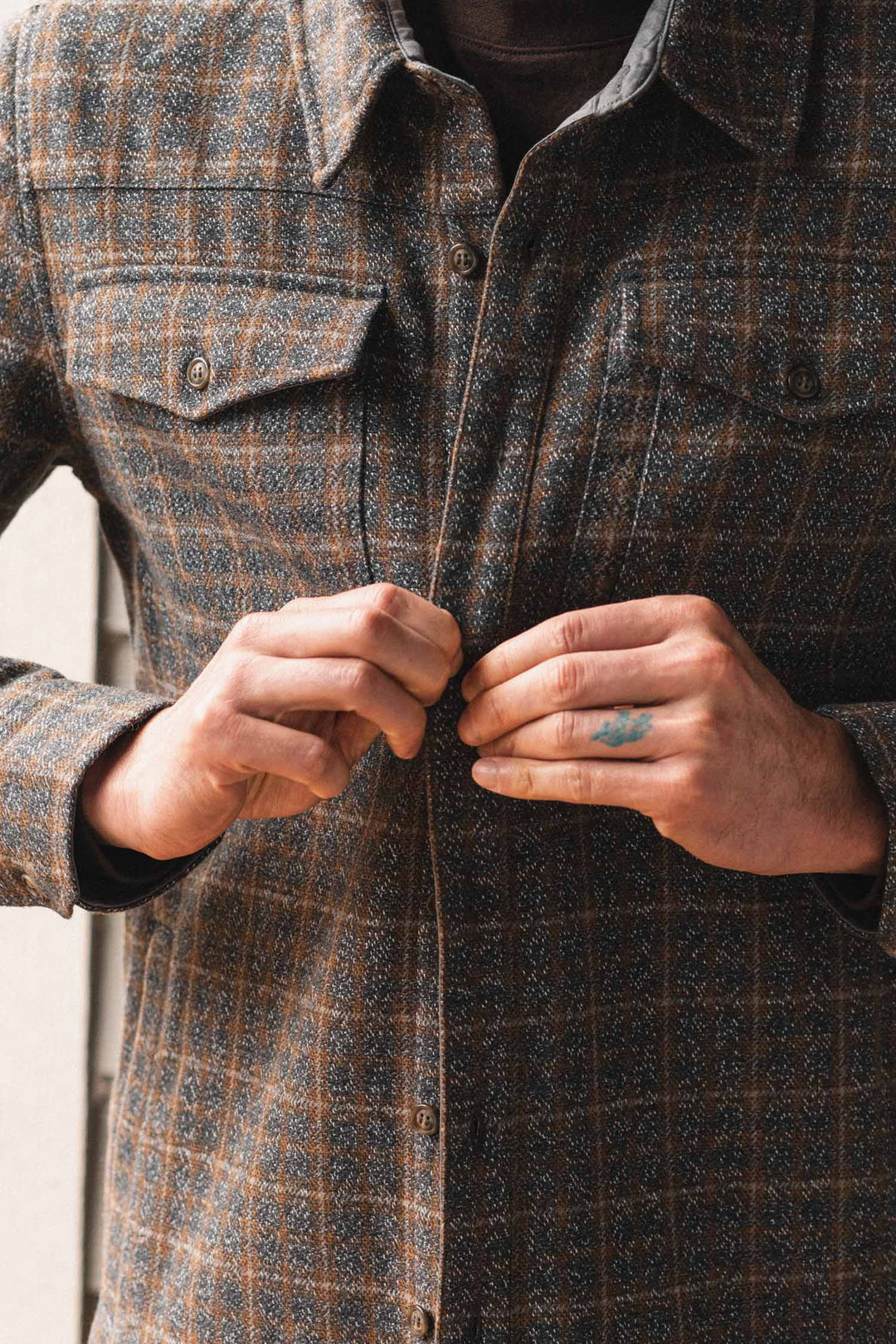 Someone buttons up the Bridge & Burn Fielding Shirt Jacket in Hearth Plaid, showing their hands—one with a small blue tattoo. The photo centers on the workwear-inspired flannel jacket and hands.