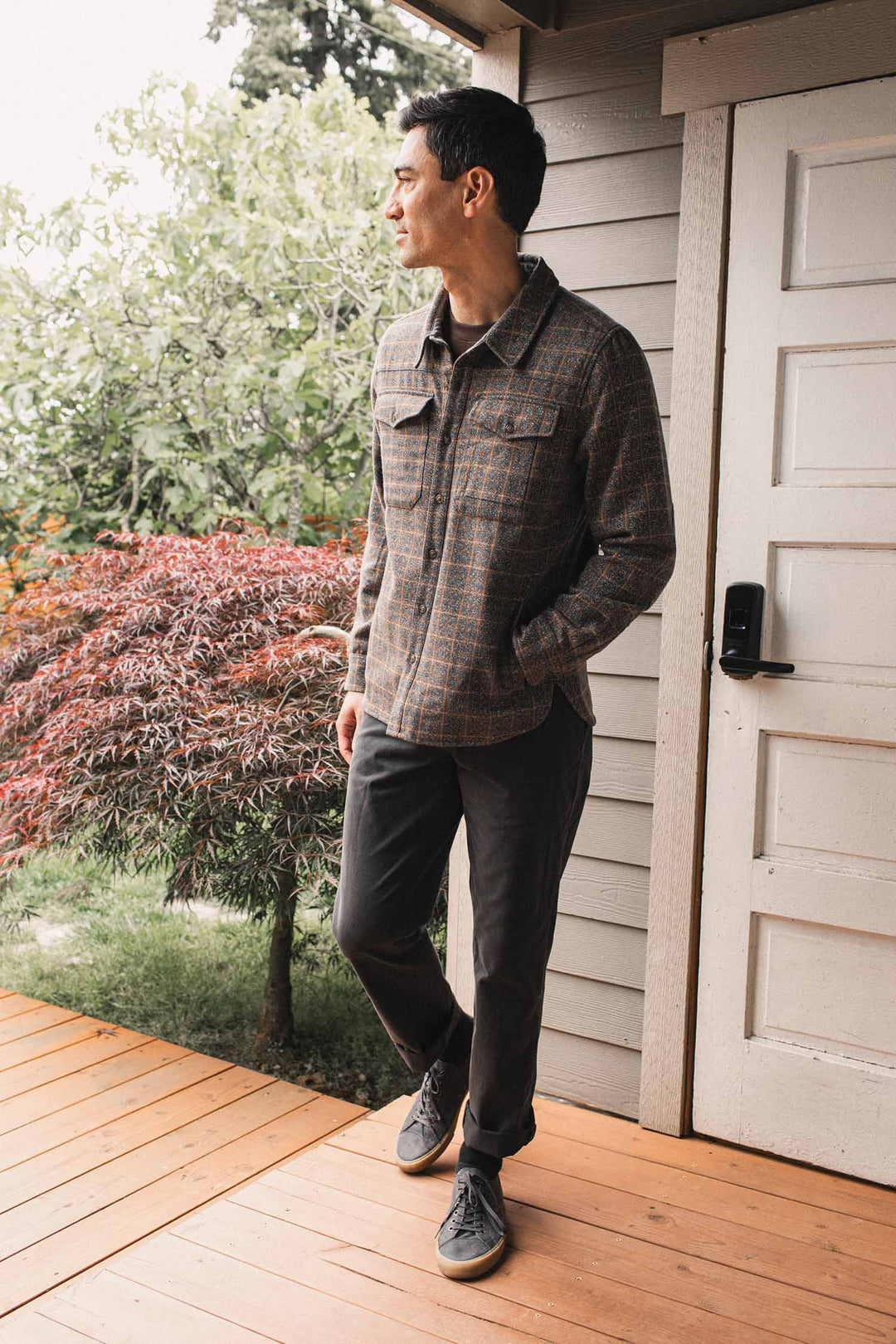 A man stands on a wooden porch near a white door, looking to the side. He wears the Bridge & Burn Fielding Shirt Jacket in Hearth Plaid, dark pants, and gray sneakers. Trees and plants are visible in the background.