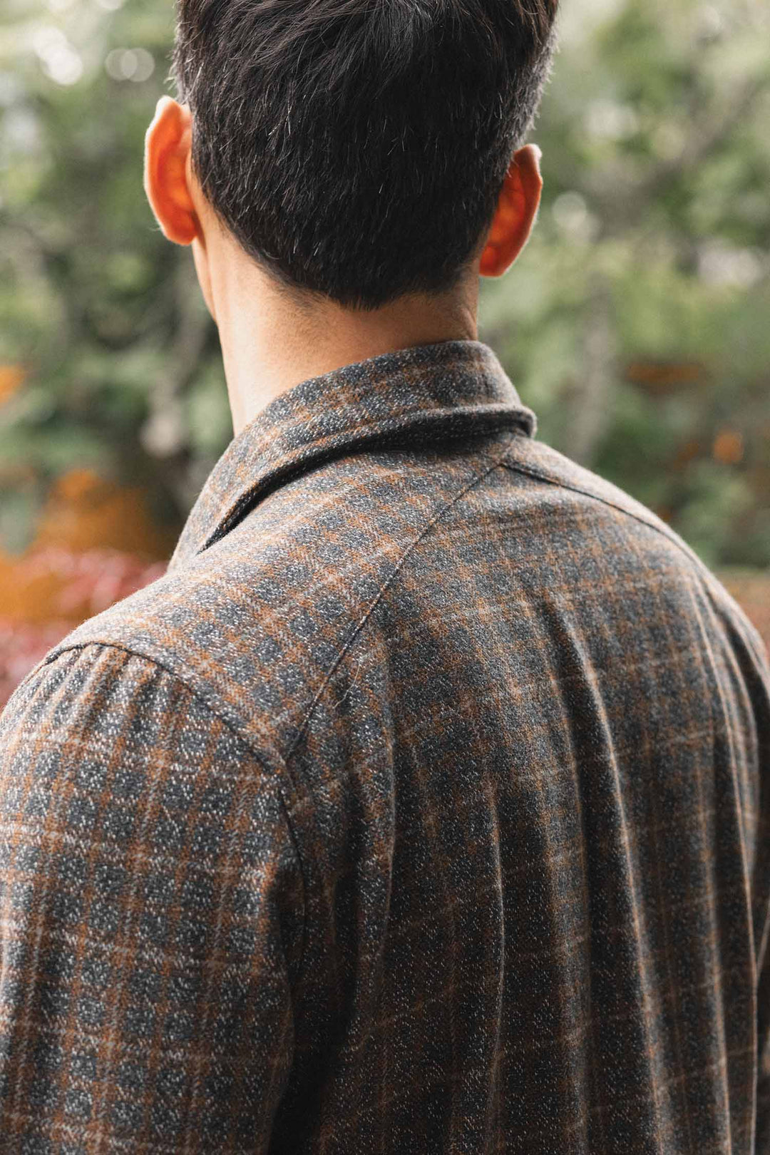 A person with short dark hair is seen from behind, wearing the Bridge & Burn Fielding Shirt Jacket in Hearth Plaid. The blurred background features green foliage with touches of orange and purple.