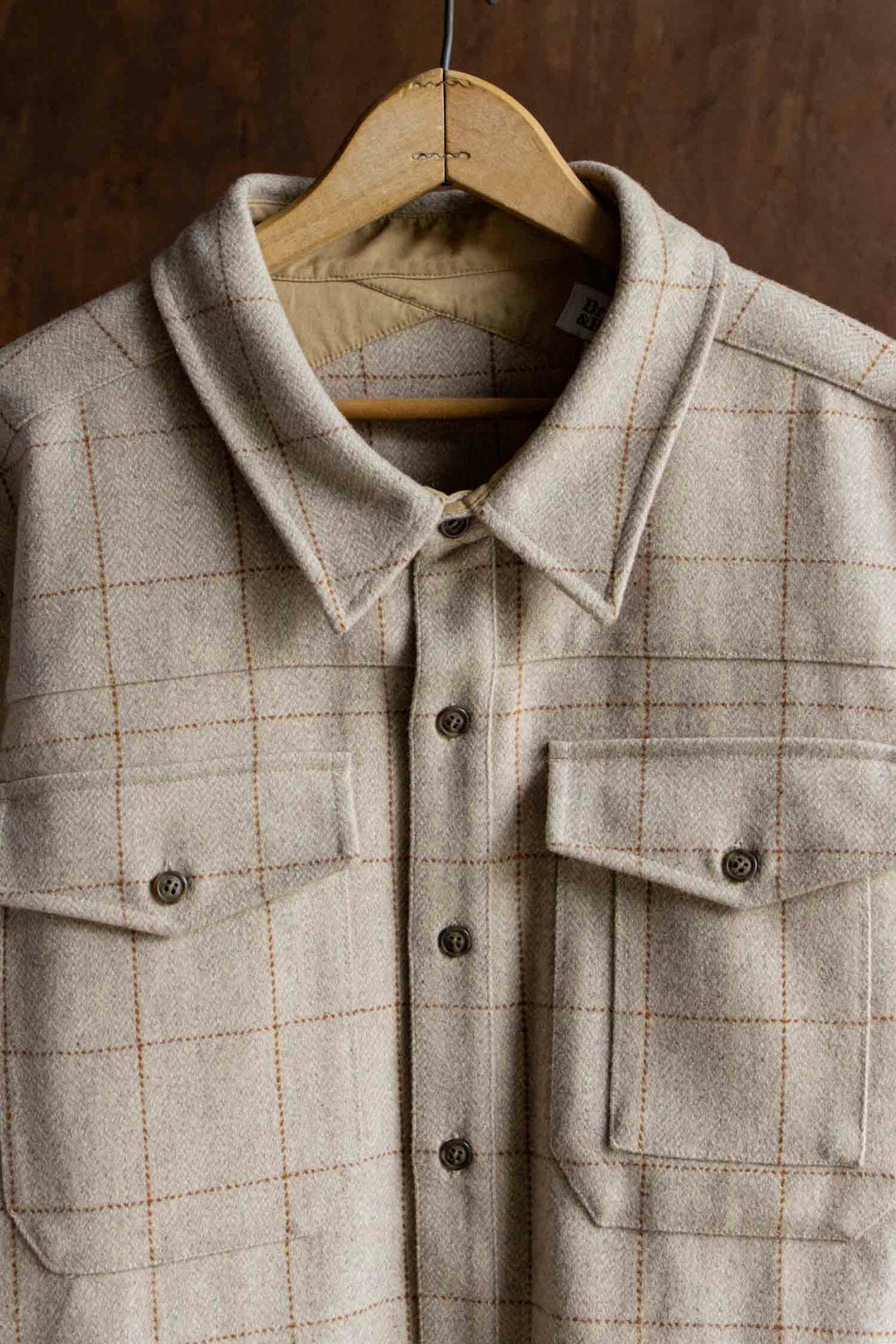 The Bridge & Burn Fielding Shirt Jacket in Stonecross, a beige plaid heavyweight overshirt with subtle orange grid lines, brown buttons, and two front pockets, is displayed on a wooden hanger against a brown background.