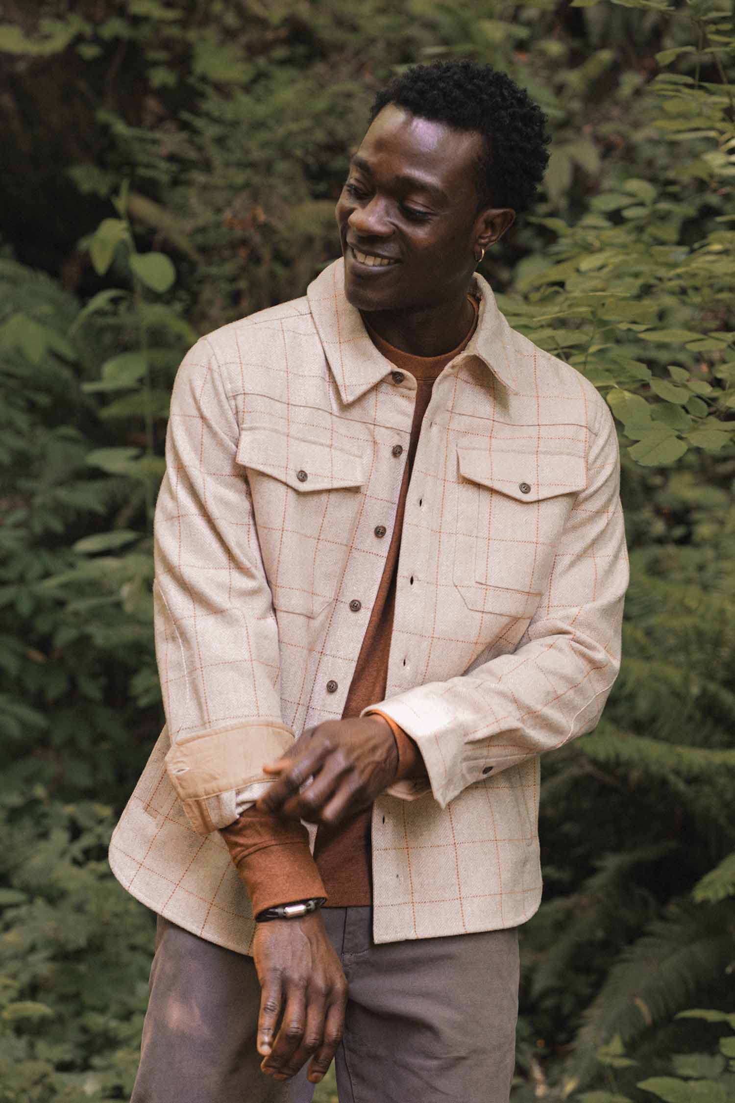A man outdoors, surrounded by greenery, smiles while rolling up the sleeve of his Bridge & Burn Fielding Shirt Jacket in Stonecross, layered over a brown shirt.