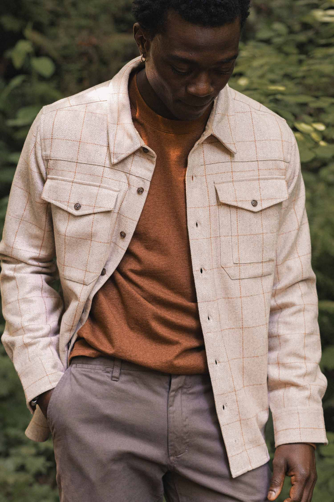 A man stands outdoors in greenery, wearing the Bridge & Burn Fielding Shirt Jacket in Stonecross over a rust shirt and gray pants, one hand in his pocket and looking down.