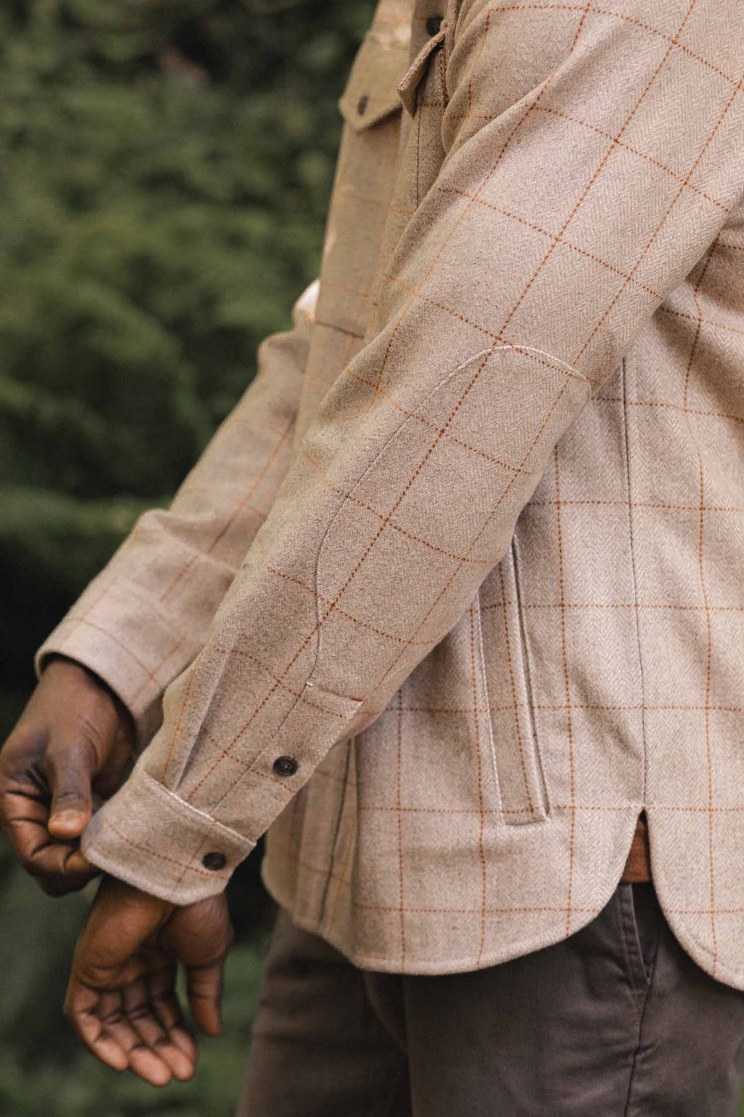 A person wearing the Bridge & Burn Fielding Shirt Jacket in Stonecross, featuring a light brown plaid pattern and brown buttons, stands outdoors with one arm slightly bent and greenery behind them.