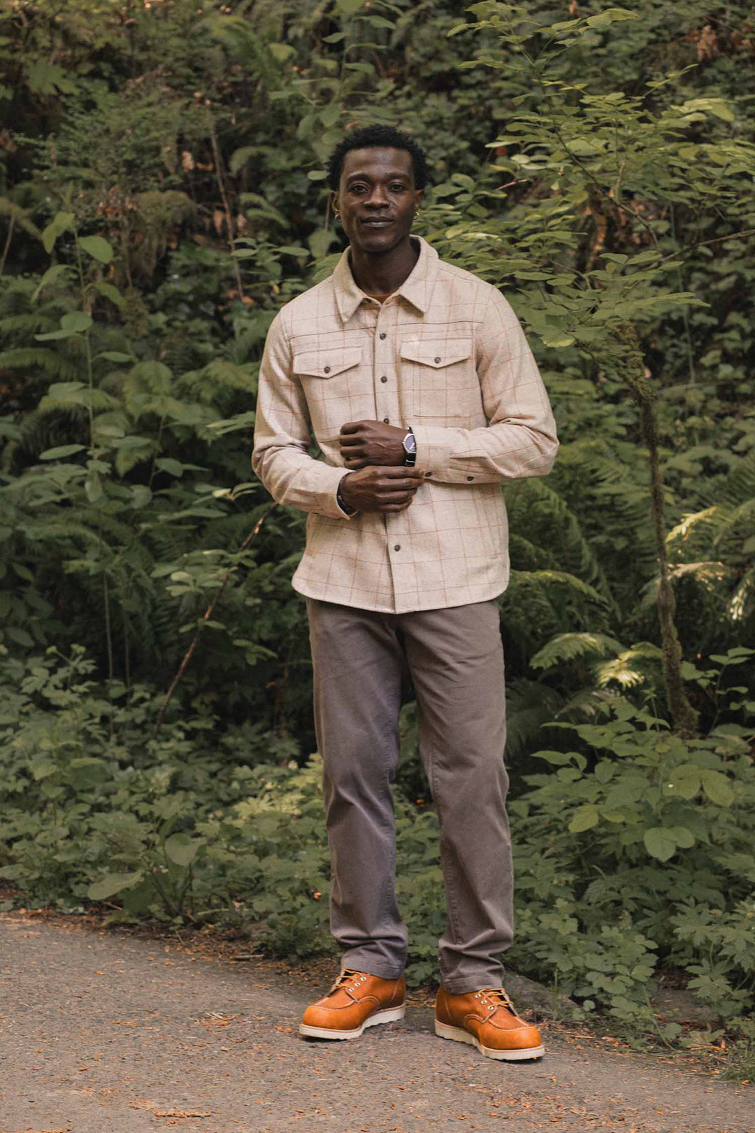 A man stands on a forest path in the Bridge & Burn Fielding Shirt Jacket in Stonecross, paired with gray pants and orange boots, surrounded by lush greenery and ferns. He smiles with his hands folded in front of him.